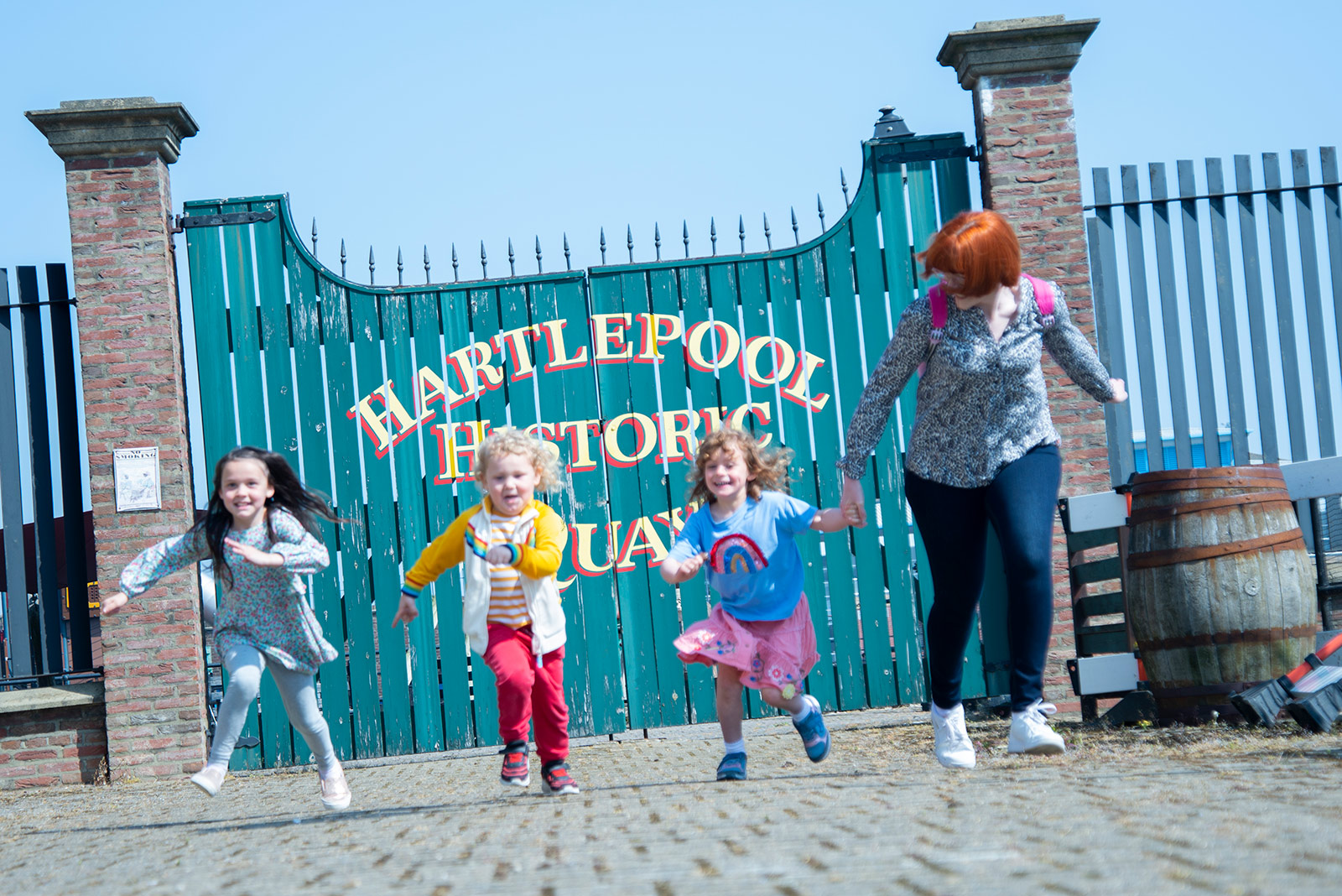 A woman with three young girls are running towards the camera. Behind them is a large blue gate that says Hartlepool Historic Quays in yellow font.