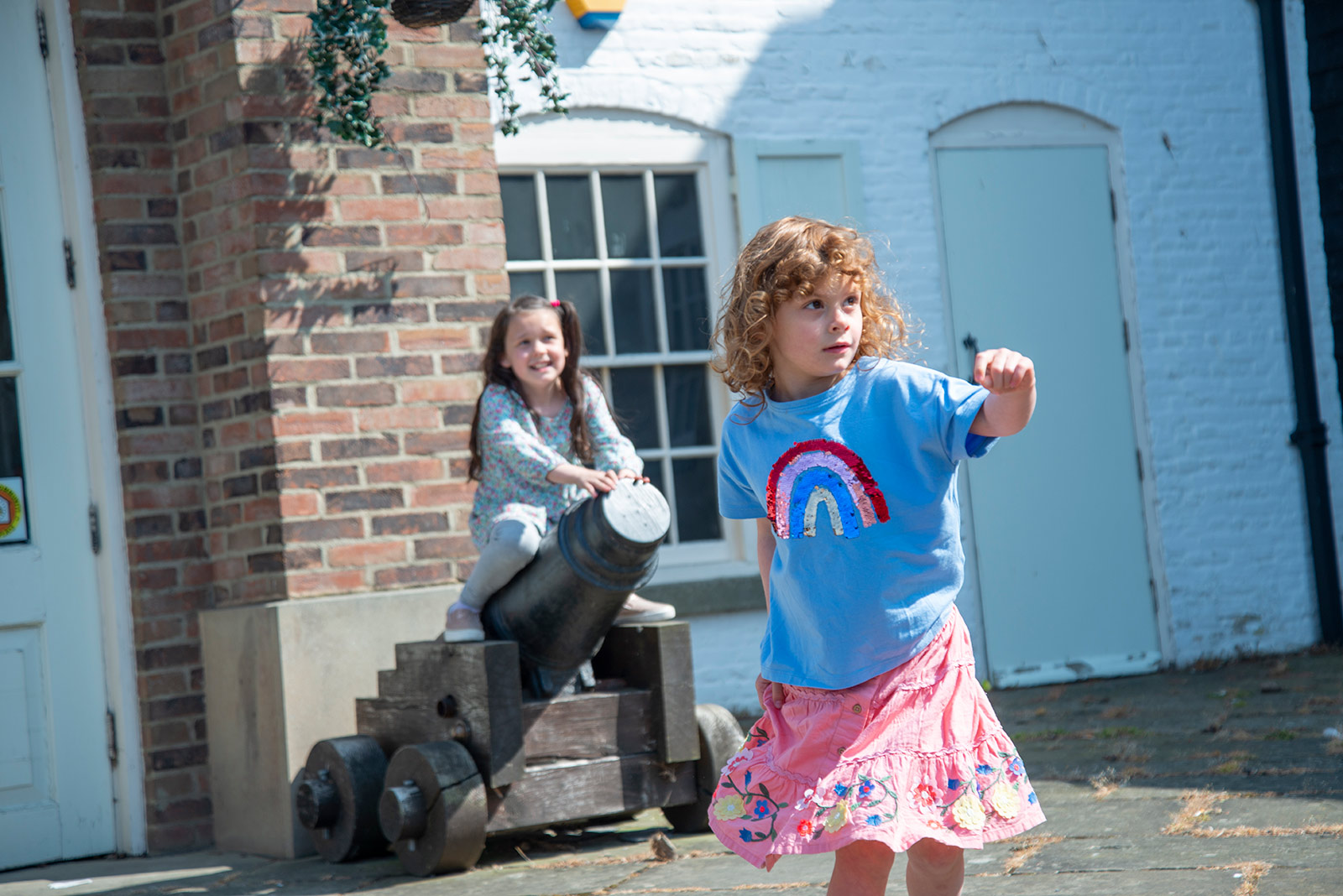 An image of two young girls in front of a brick building. One of the girls is sitting on top of a small canon.