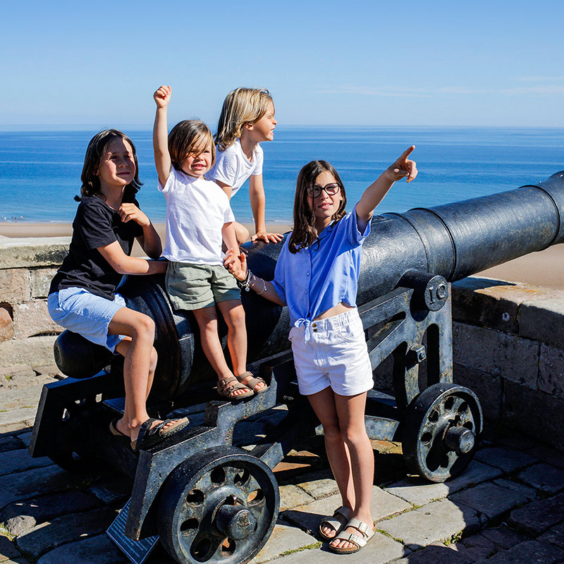 A group of four children sit on top of a cannon over looking the sea.