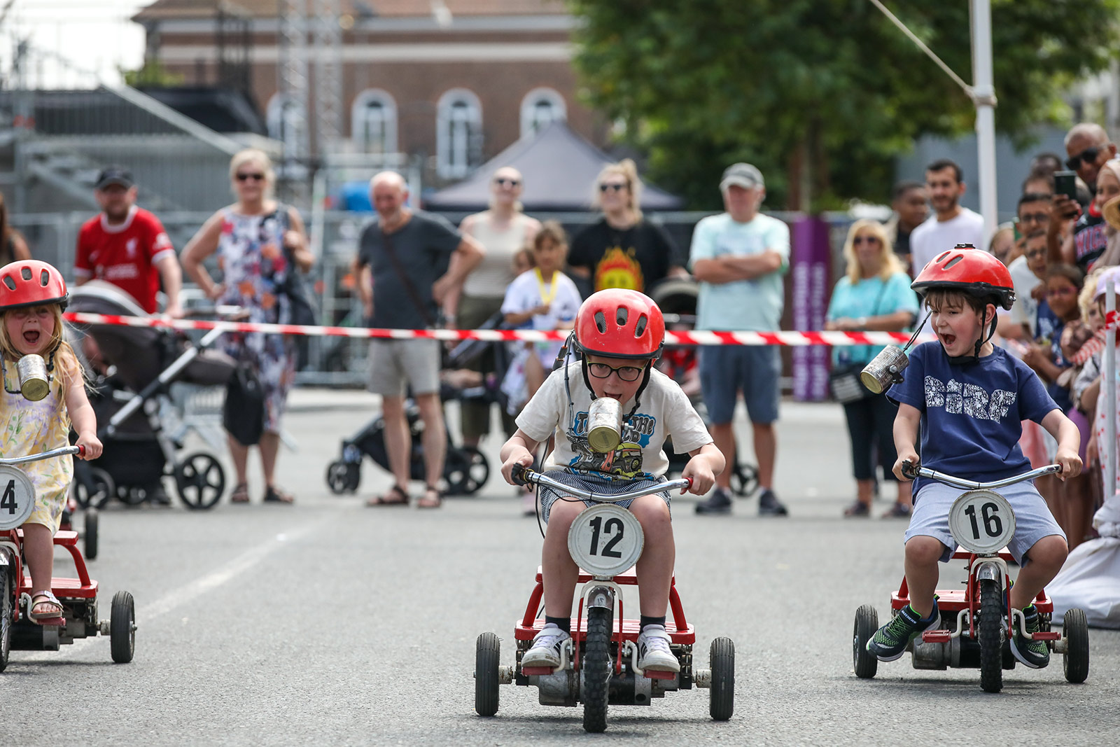 Children race down a street on trikes and wearing red helmets. In the background there are grown ups watching behind a red and white tape.