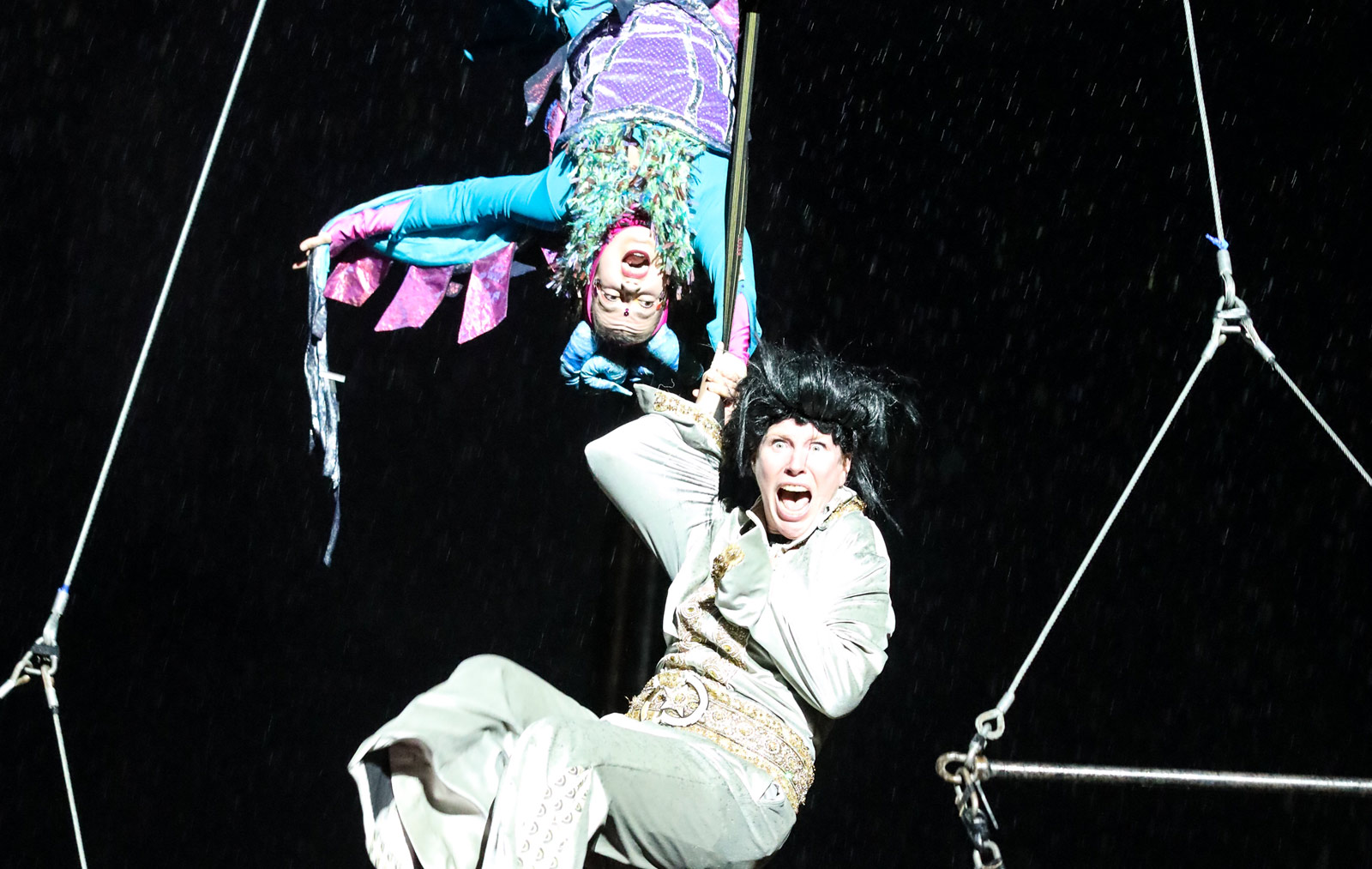 Two acrobats hang from an Ariel swing against a black background.