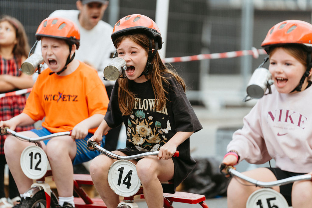 Three children are lined up at a starting line on small trikes each with numbers on. They are wearing orange helmets and look excited.