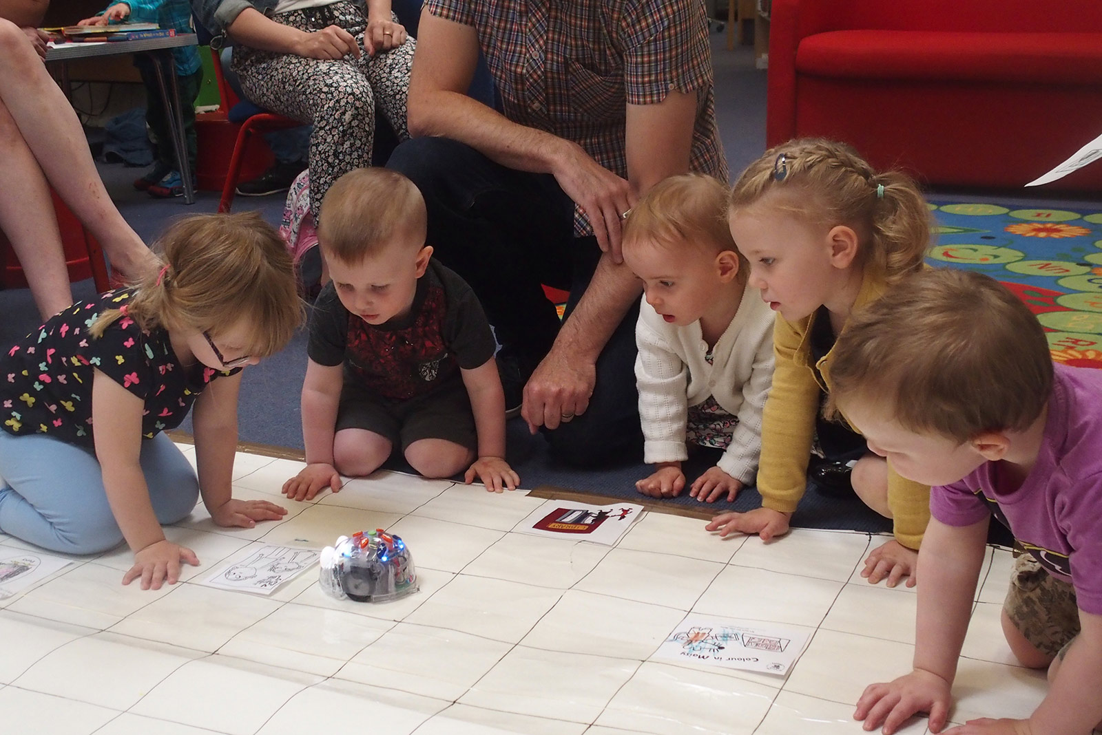 A group of young children sit around white paper squares watching a beebot light up.