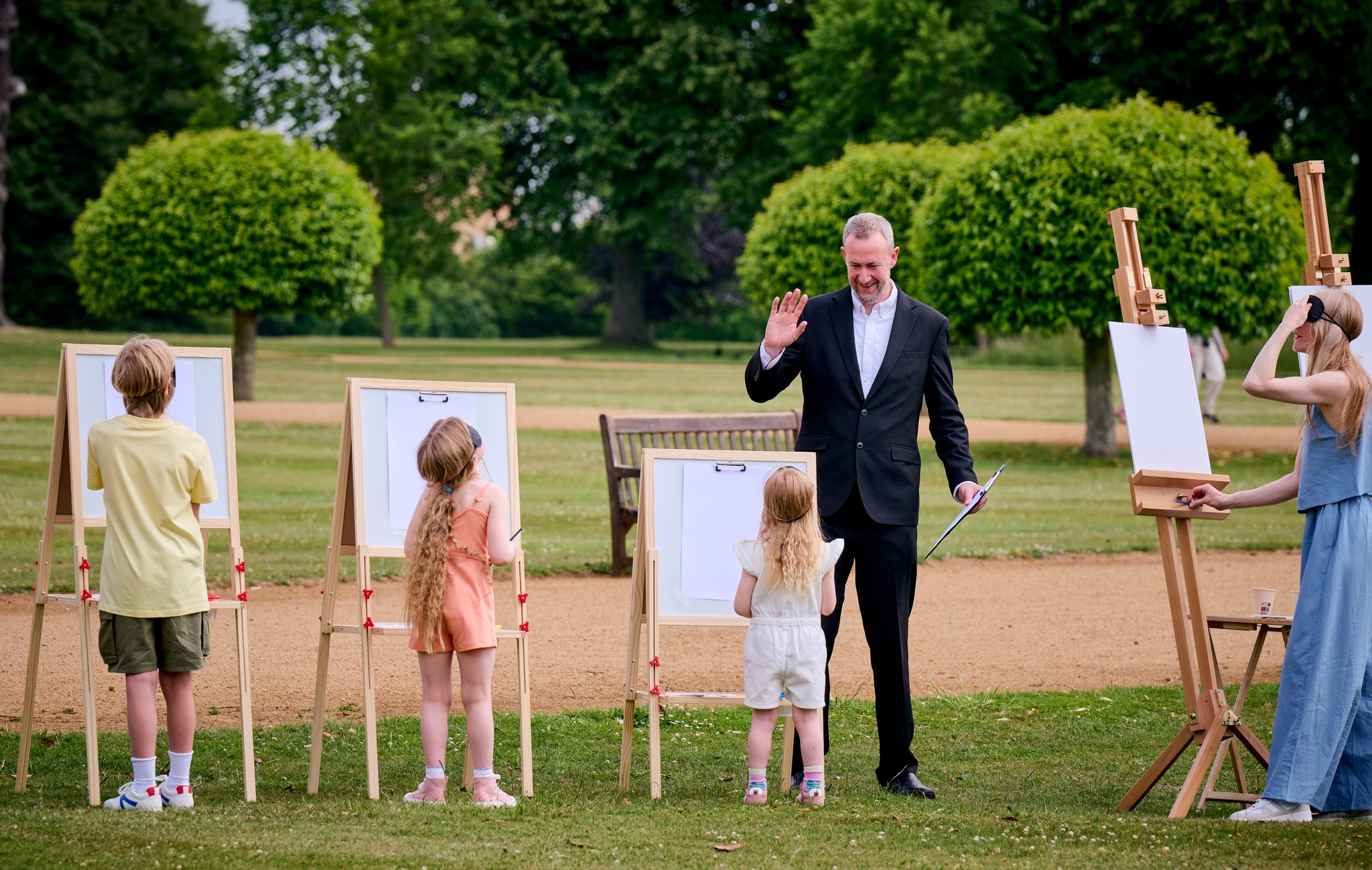 A group of children stand in front of easels with their backs to the camera. A man in a suit is leading an activity in an outdoor setting.