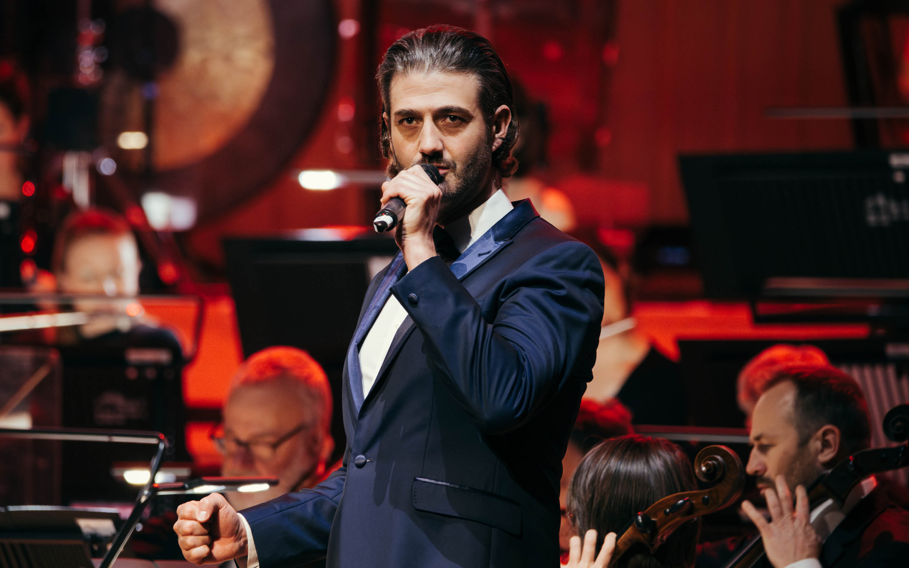 A man in a suit sings into a microphone. An orchestra can be seen behind.