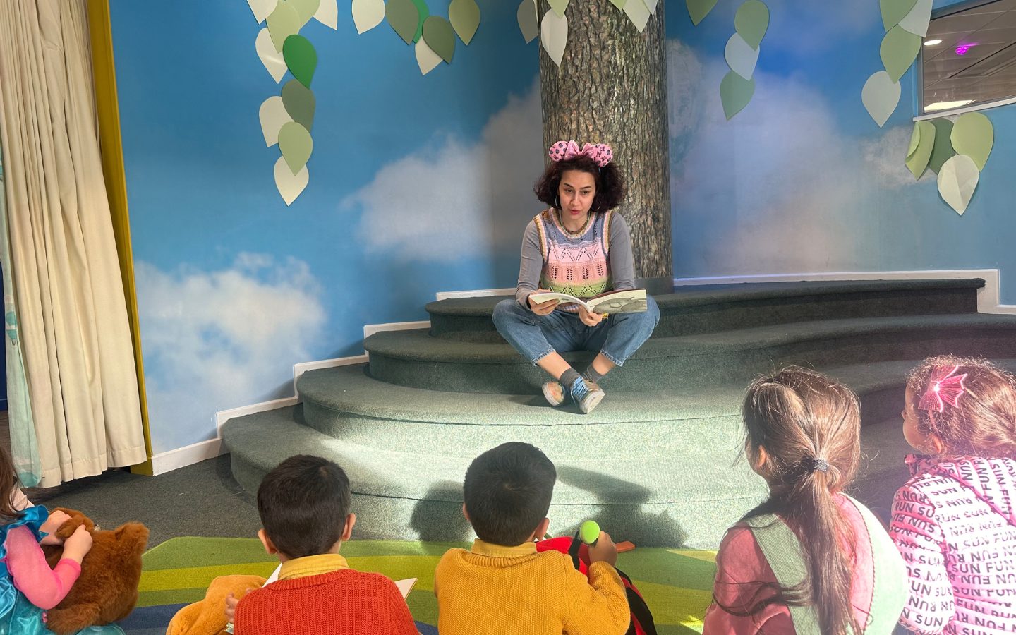 An adult with a big pink bow in their hair sits indoors on some steps with a model of a tree behind them. She is reading a book t children who are looking on.