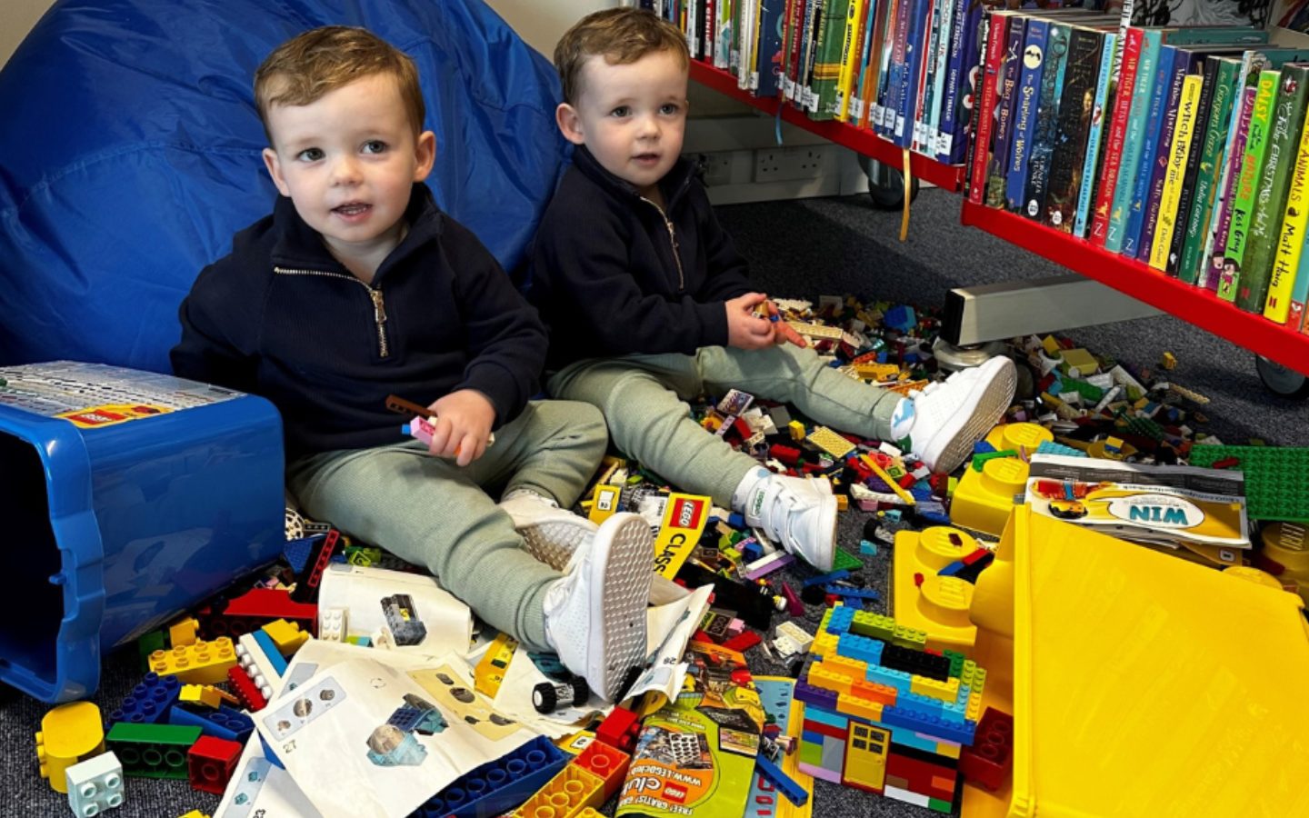 Two young children sit among lots of Lego next to a bookshelf.