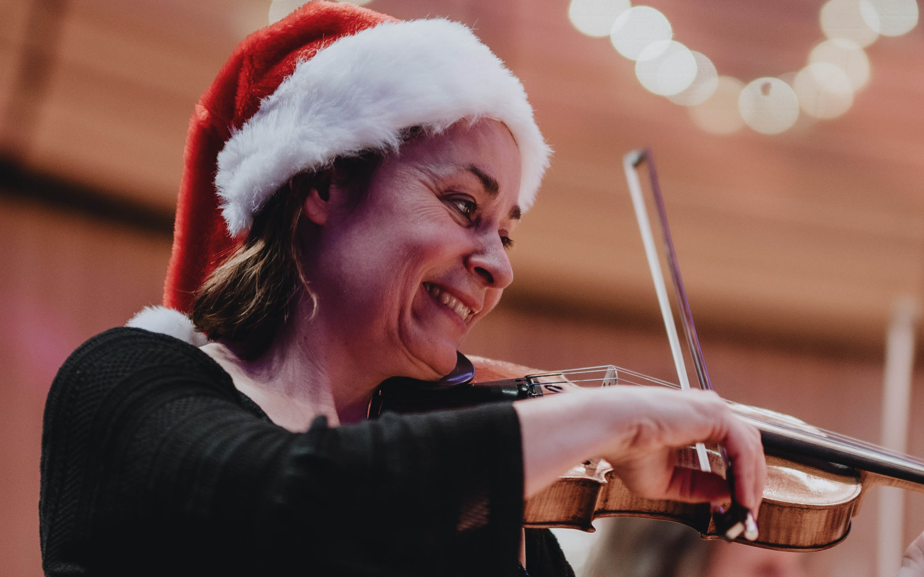 A woman in a Santa hat plays the violin, smiling
