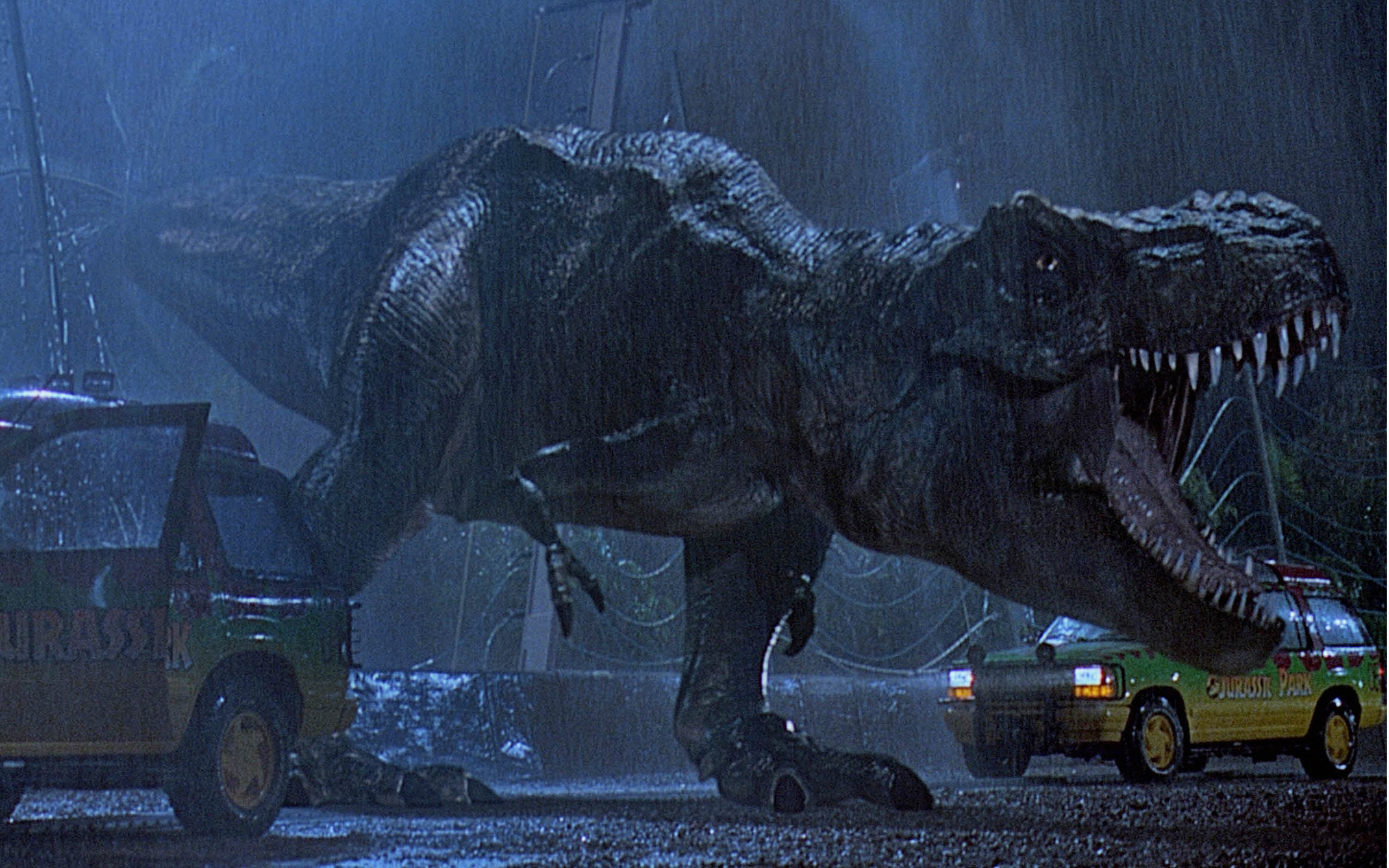 A still from a film shwing a T Rex roaring. There are vehicles around it.