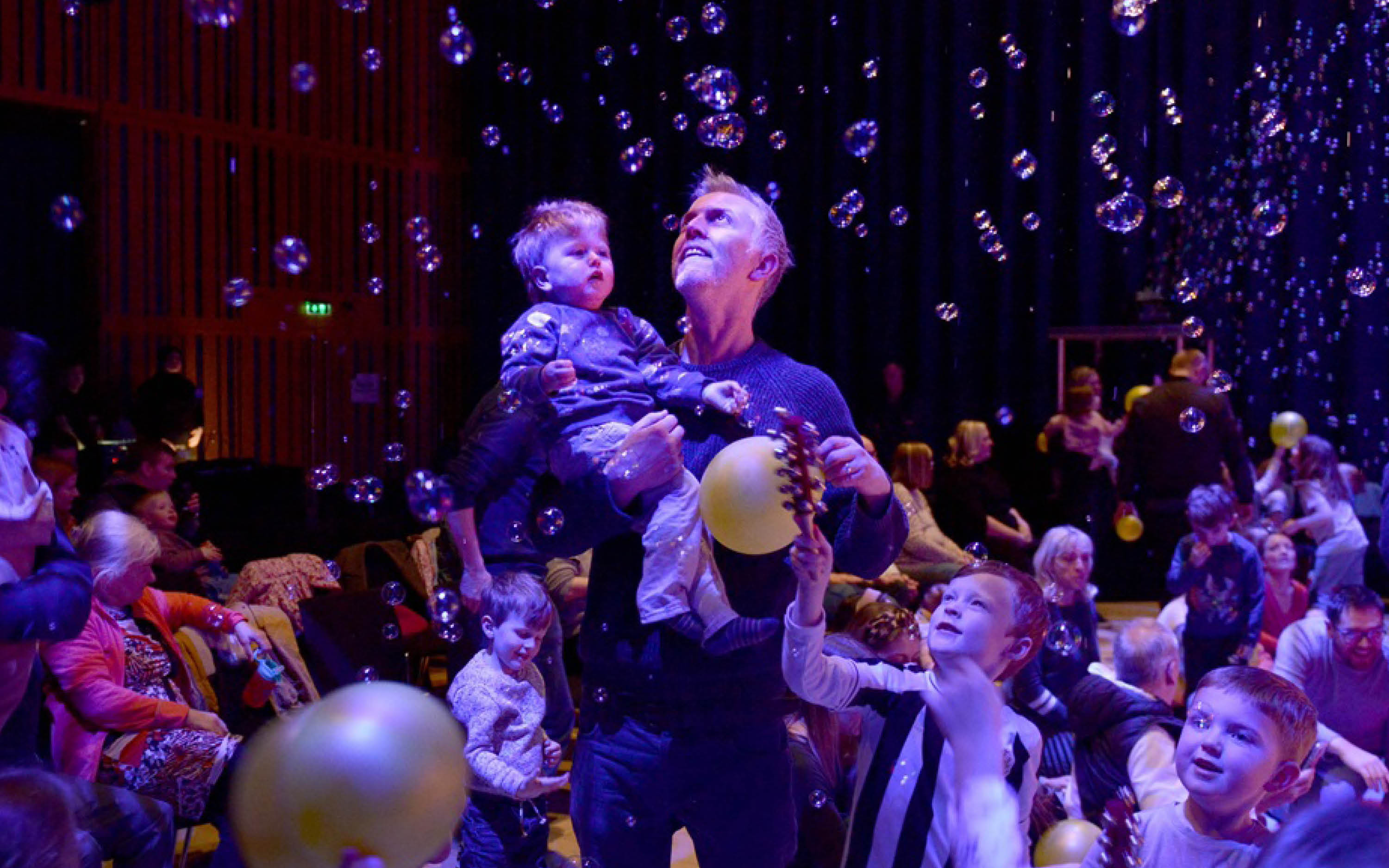 An adult carries a child. They are holding a balloon and there are bubbles raining down on them.