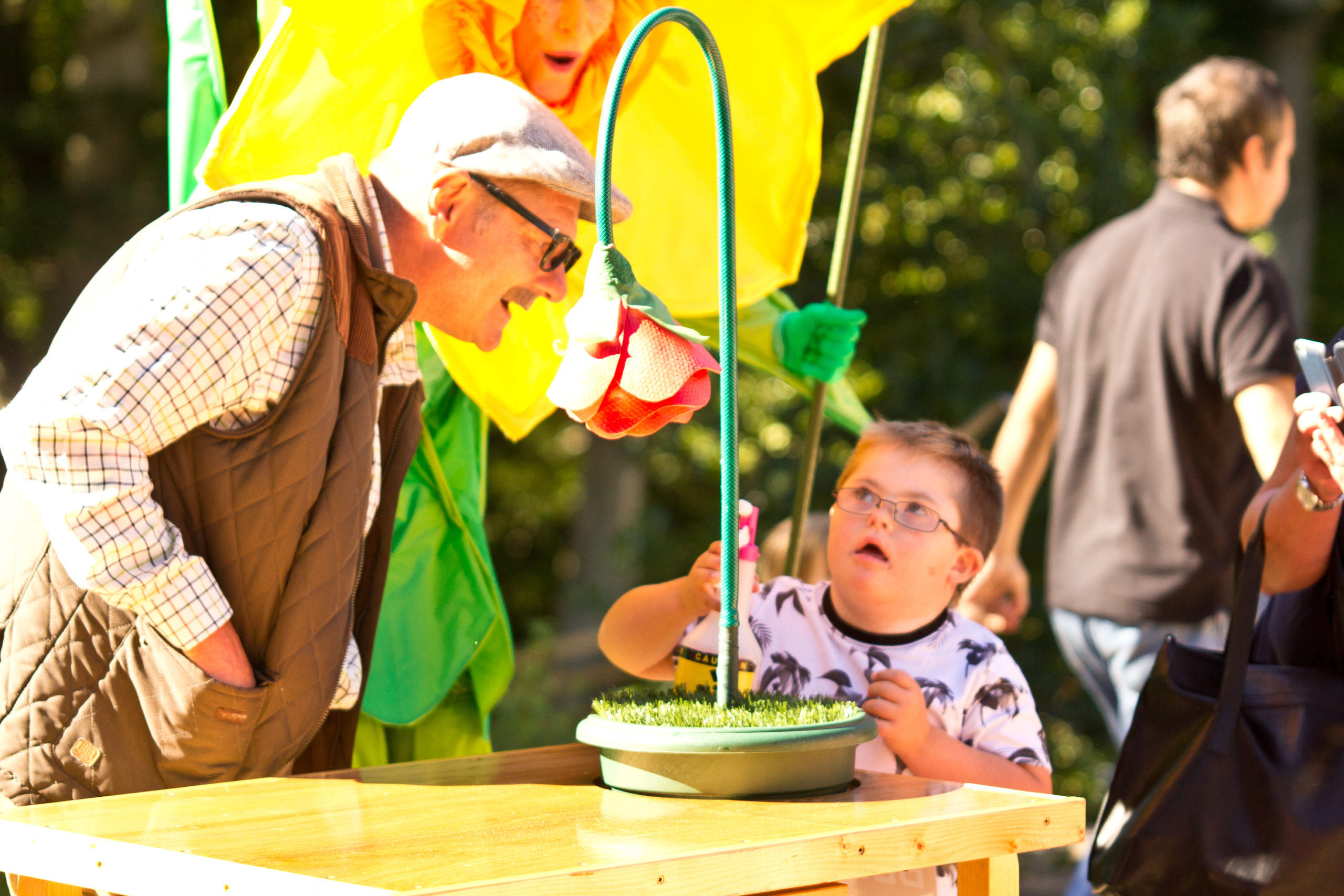 An elderly gentleman watches over a young boy as he waters a big pretend flower.