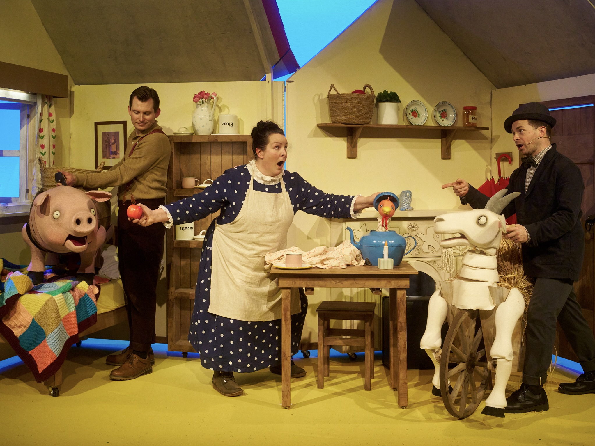 A group of actors stand on stage in a kitchen with a puppet of a pig, goat and a chicken.