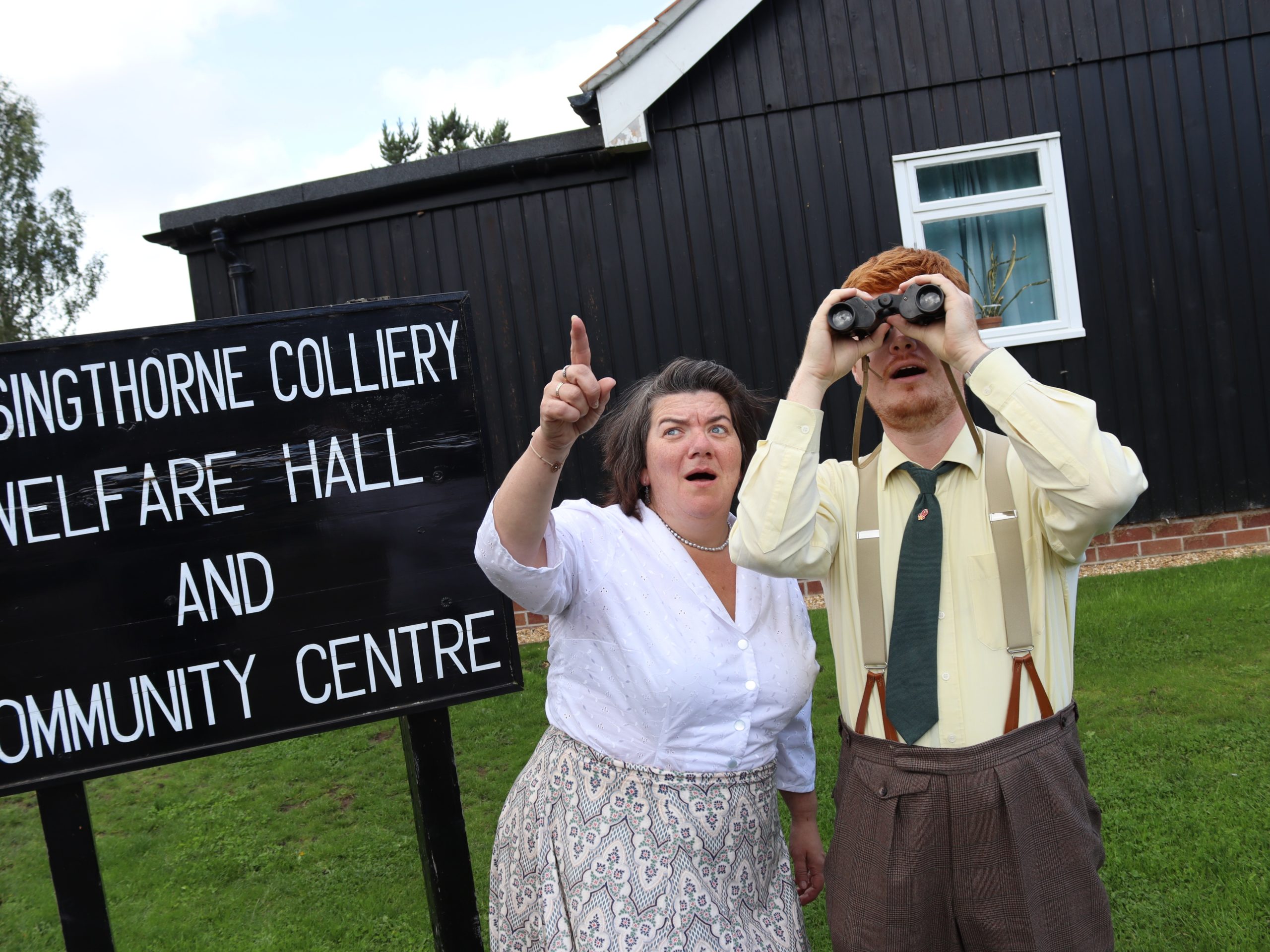 Two people look up to the sky, a man and woman wearing smart clothes. The man is looking through binoculars and the woman is pointing in amazement.