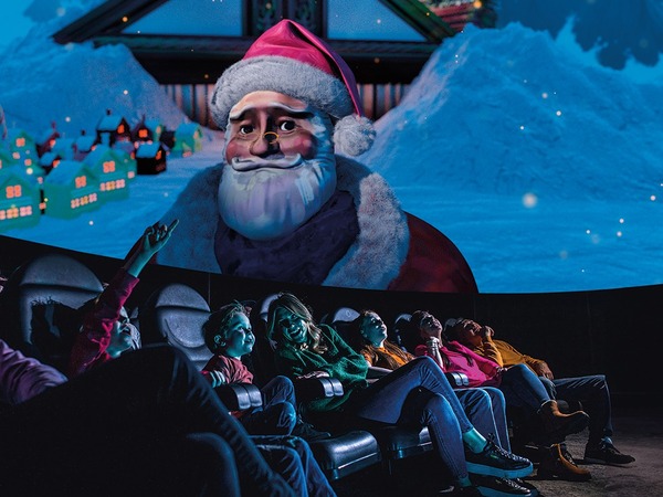 A family group sit in a planetarium looking up at the ceiling where a large Santa is above them.