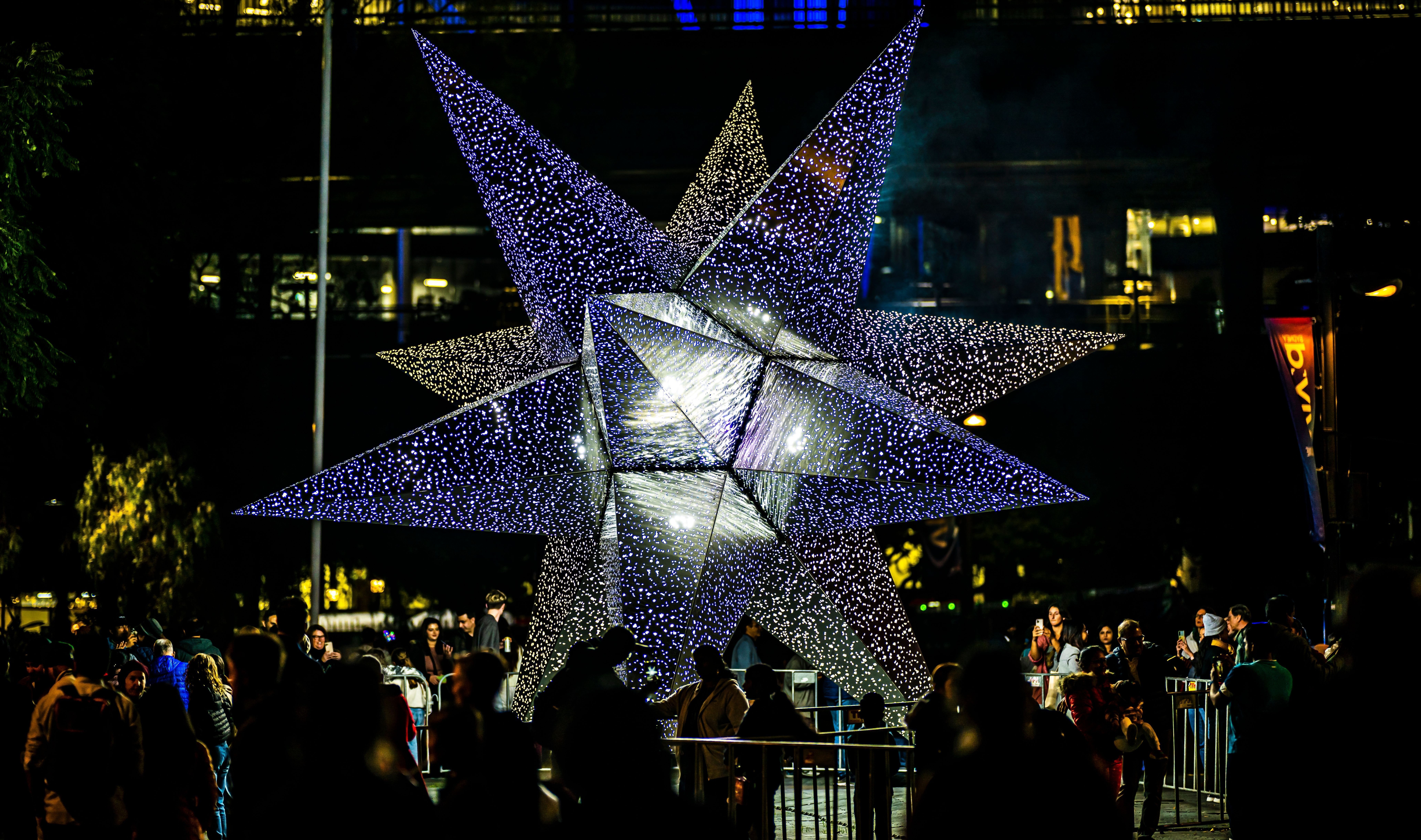 A large star shape is lit up in a city centre with a crowd of people stood looking at it.
