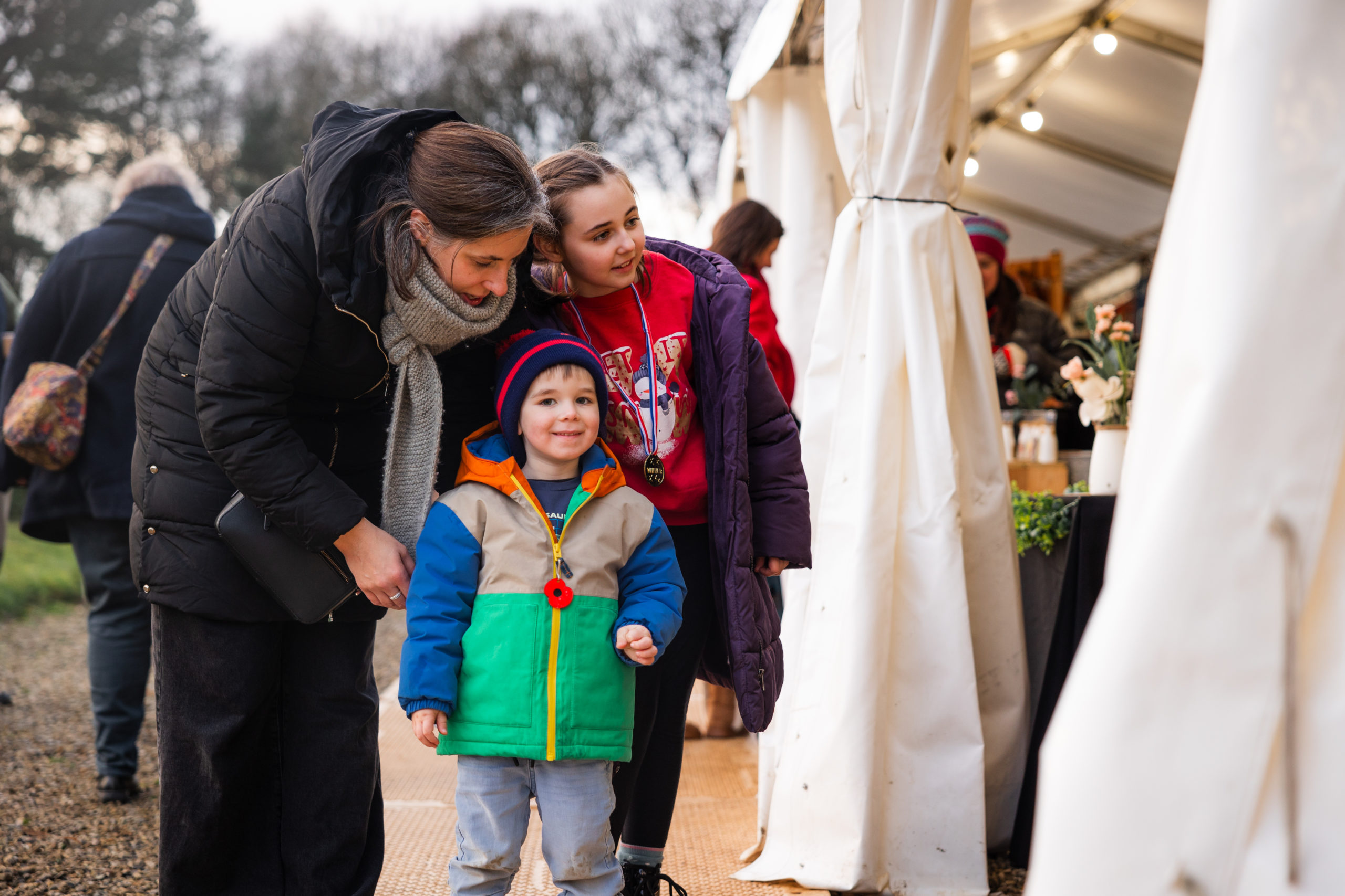 A woman stands with two children wearing coats and looking into an outdoor marquee at a winter market.