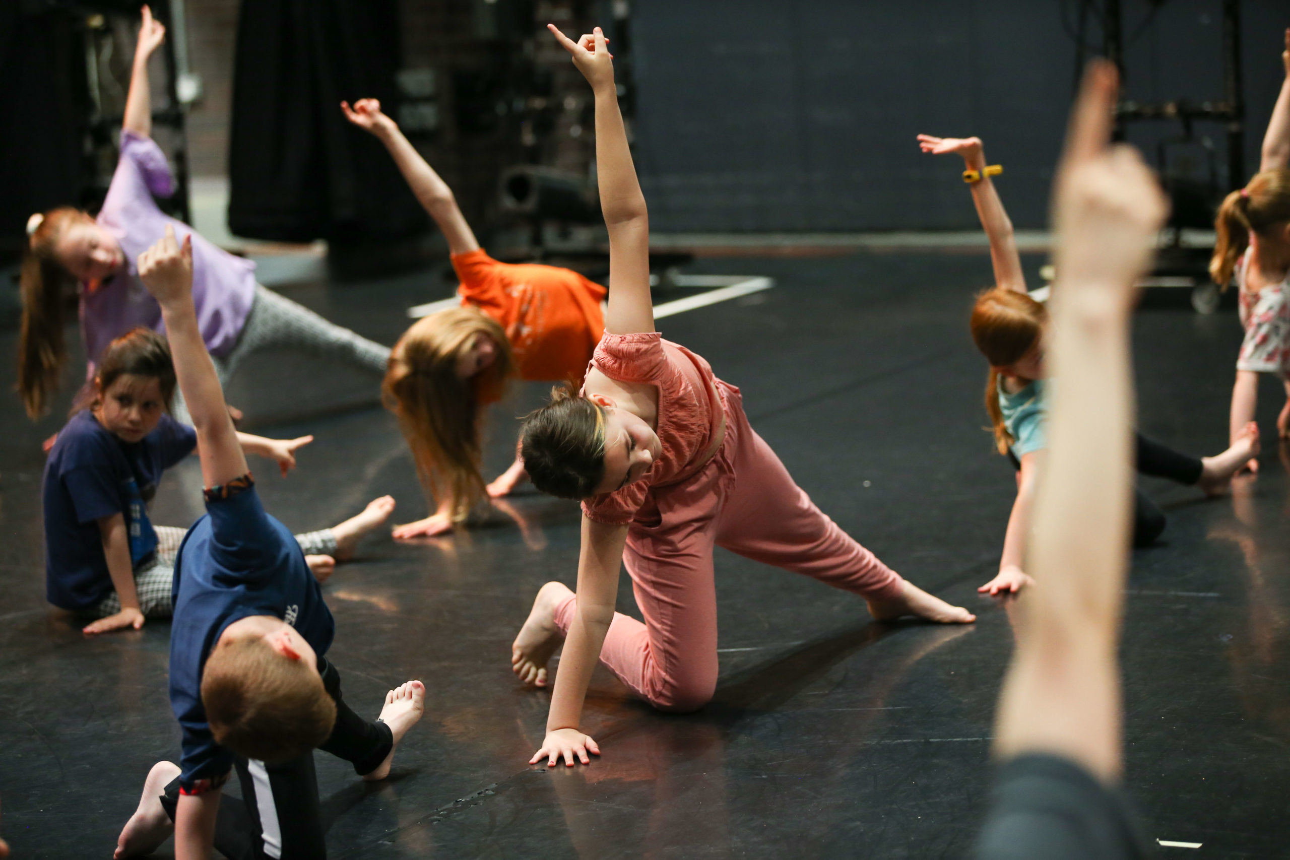 A group of children, boys and girls, stretch in a dance studio with a black floor and walls.