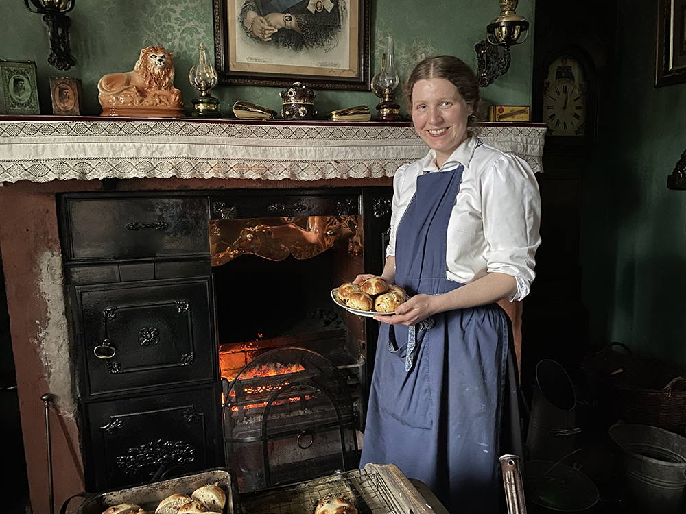 A woman stands in front of an open fire holding plate of hot cross buns. She is wearing a blue apron on top of a white shirt.