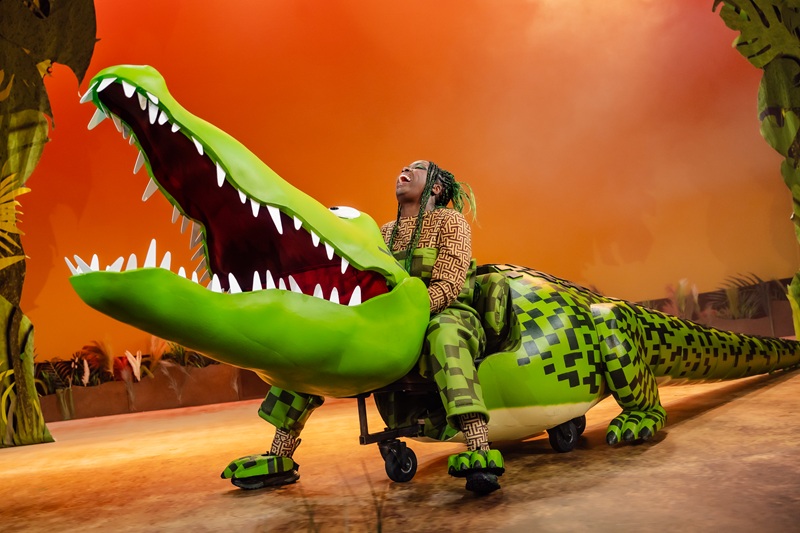 A woman sits on top of large puppet crocodile on stage in front of an orange background.