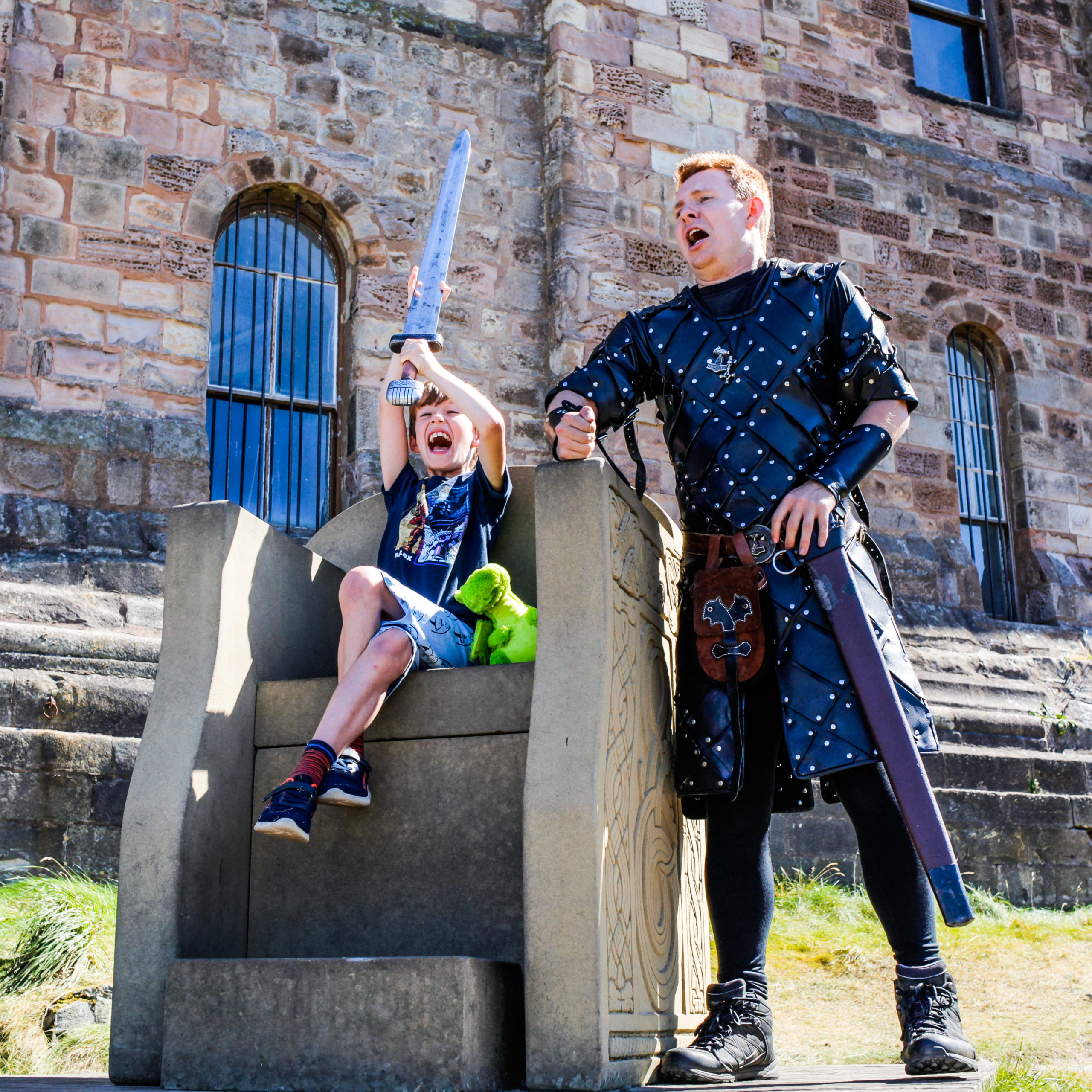 A young boy sits on a stone throne outside of a castle, holding a toy sword above his head. A man stands beside him dressed as a medieval knight.