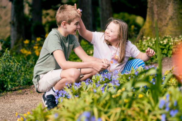 A young boy and girl crouch beside a bed of blue flowers. The girl holds one of the blue flowers up to the boys head.