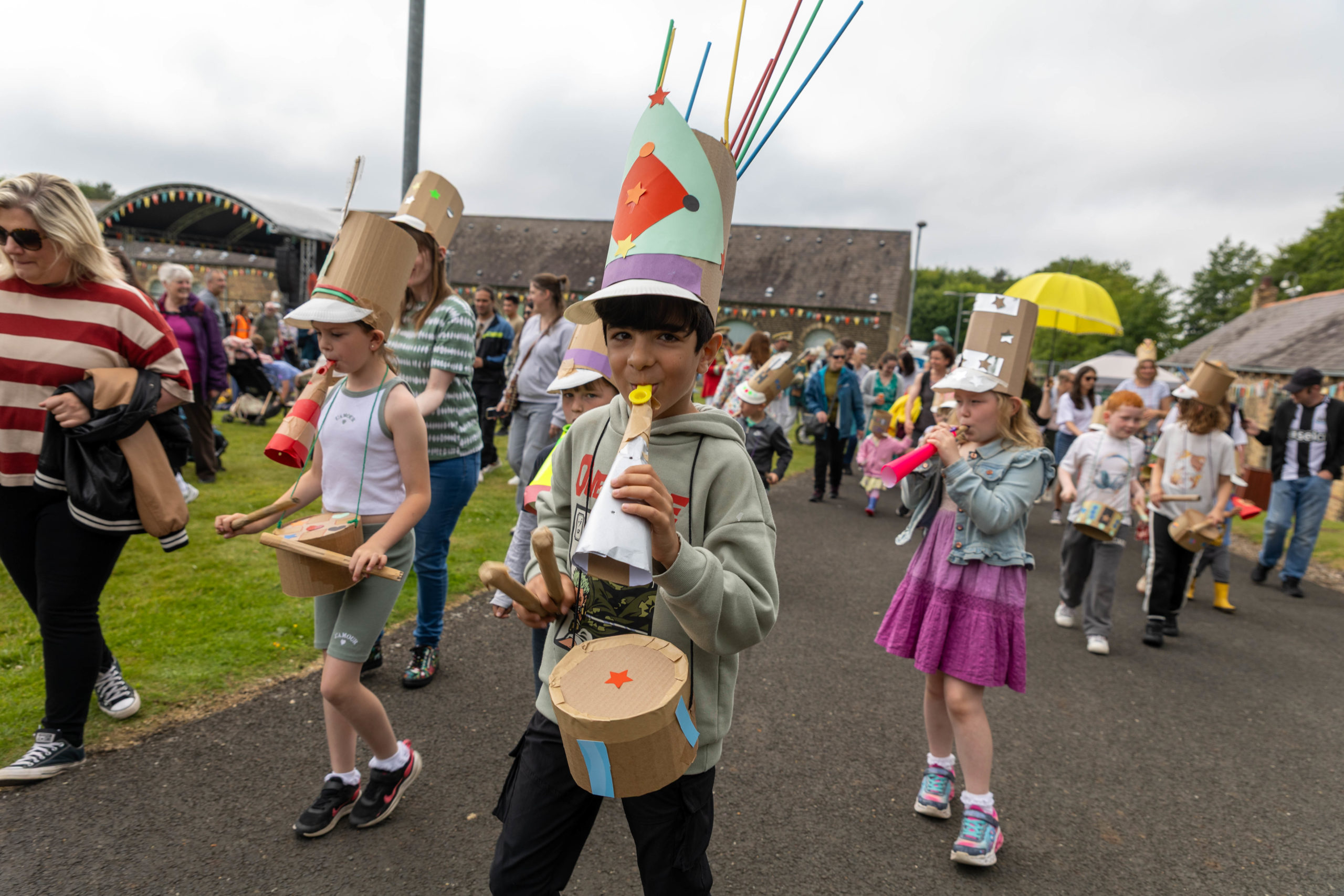 A large group of children and adults parade wearing hats and banging handmade drums.