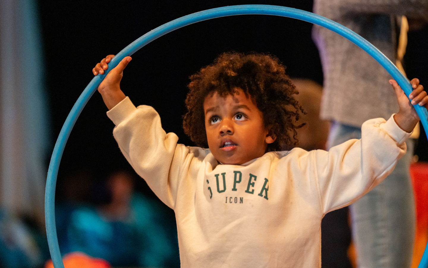 A small boy wearing a cream sweatshirt holds a blue hoola hoop ring over his head. He is looking up at it.
