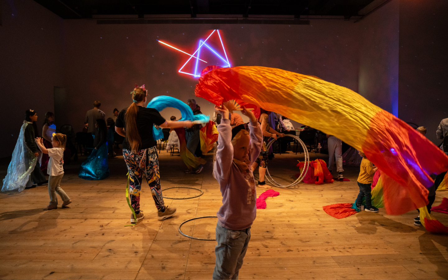 A room lit with neon lights and children inside playing with colourful materials and big hoops, some of which are lying on the wooden floor.