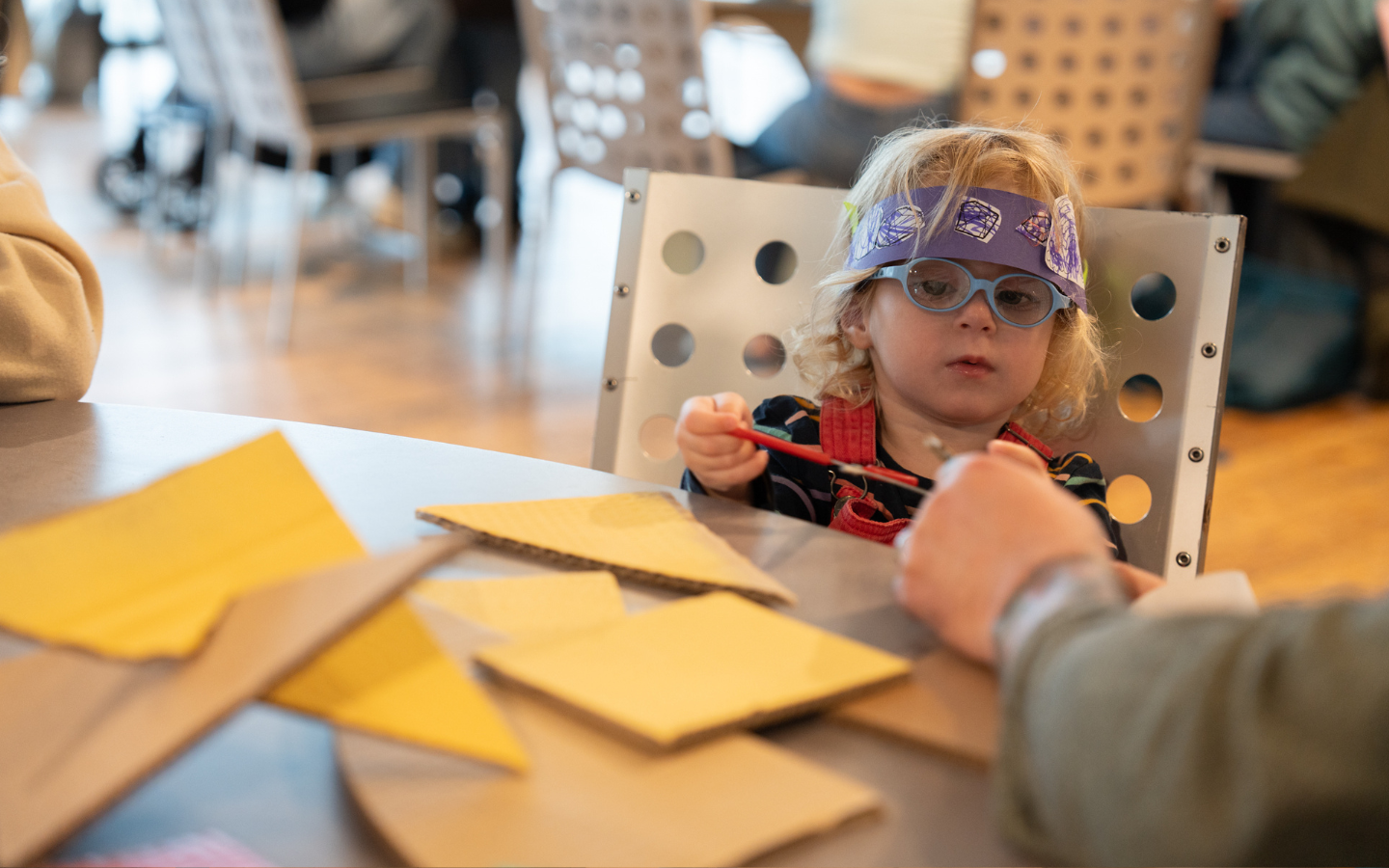 An image of a small child sitting at a craft table snipping with a pair of red handled scissors. There is yellow card on the table cut into triangles and an adult is helping the child cut.