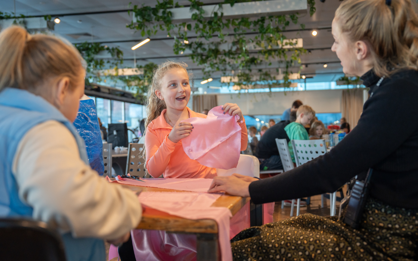A family group sit at a table with pink material laid out in front of them. One girl holds up a piece of the pink material that has been cut into a heart shape.