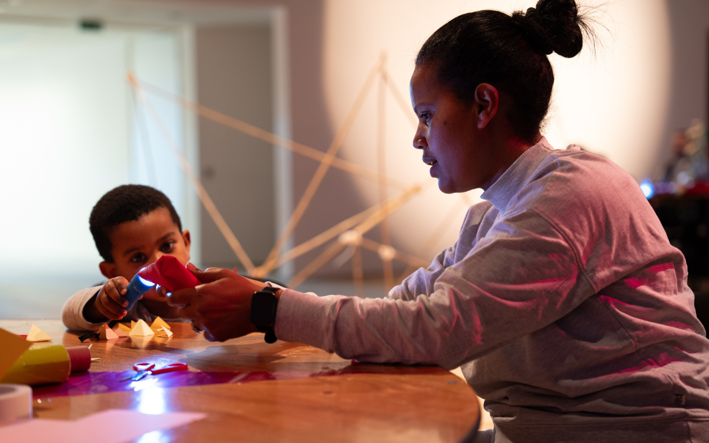 An image of a lady leading an activity session sitting at a table with a young boy. They are shining a light on to the table top.