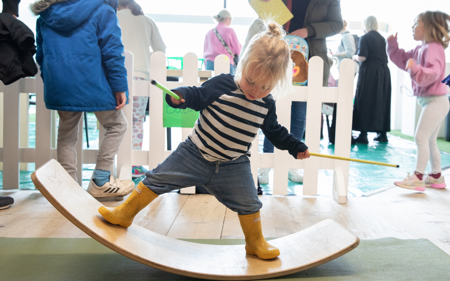 A young girl balances on a curved wooden beam and holds a yellow and green button in each hand.