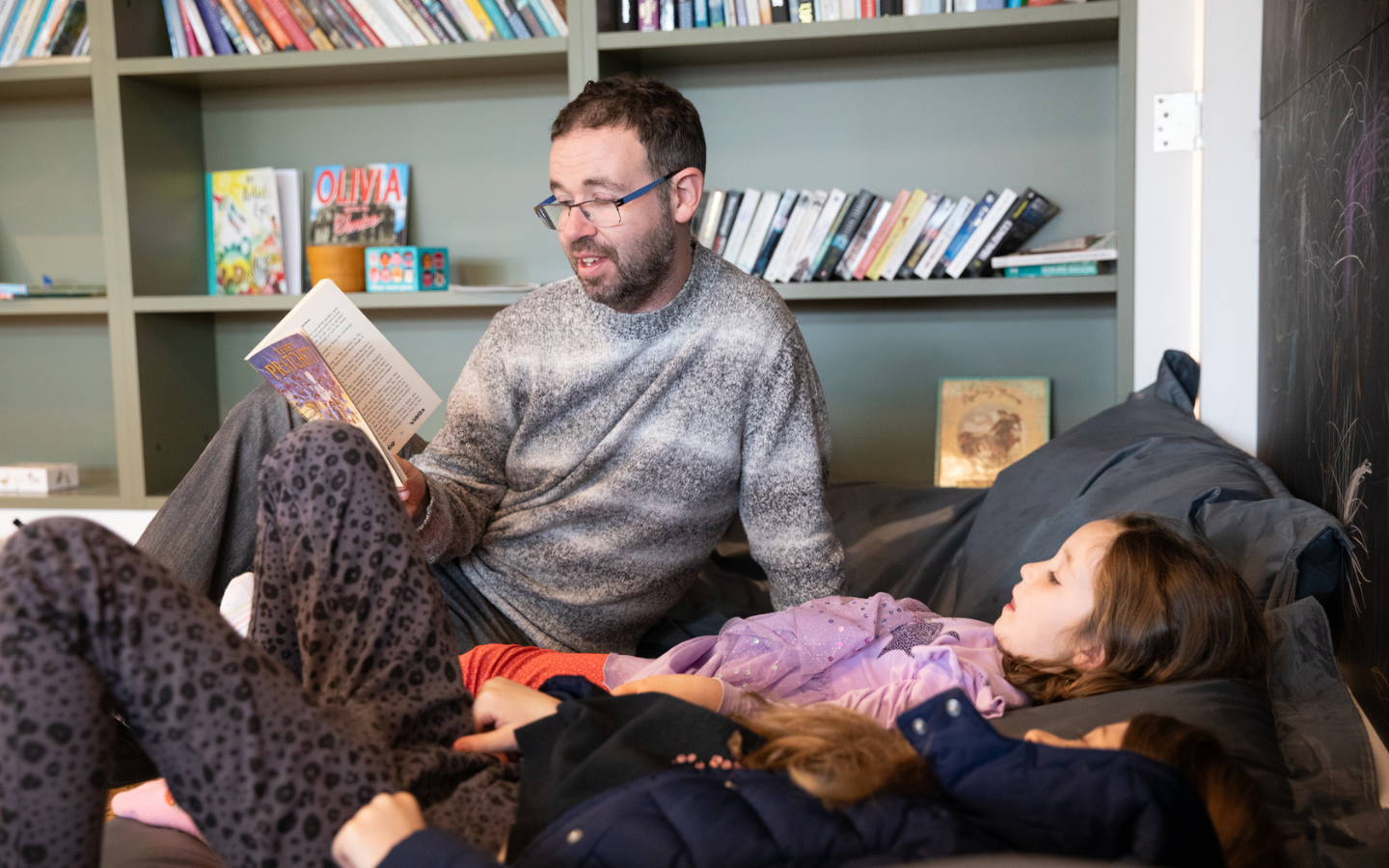 A man and a child sit on beanbags reading together. In the background is a bookshelf.