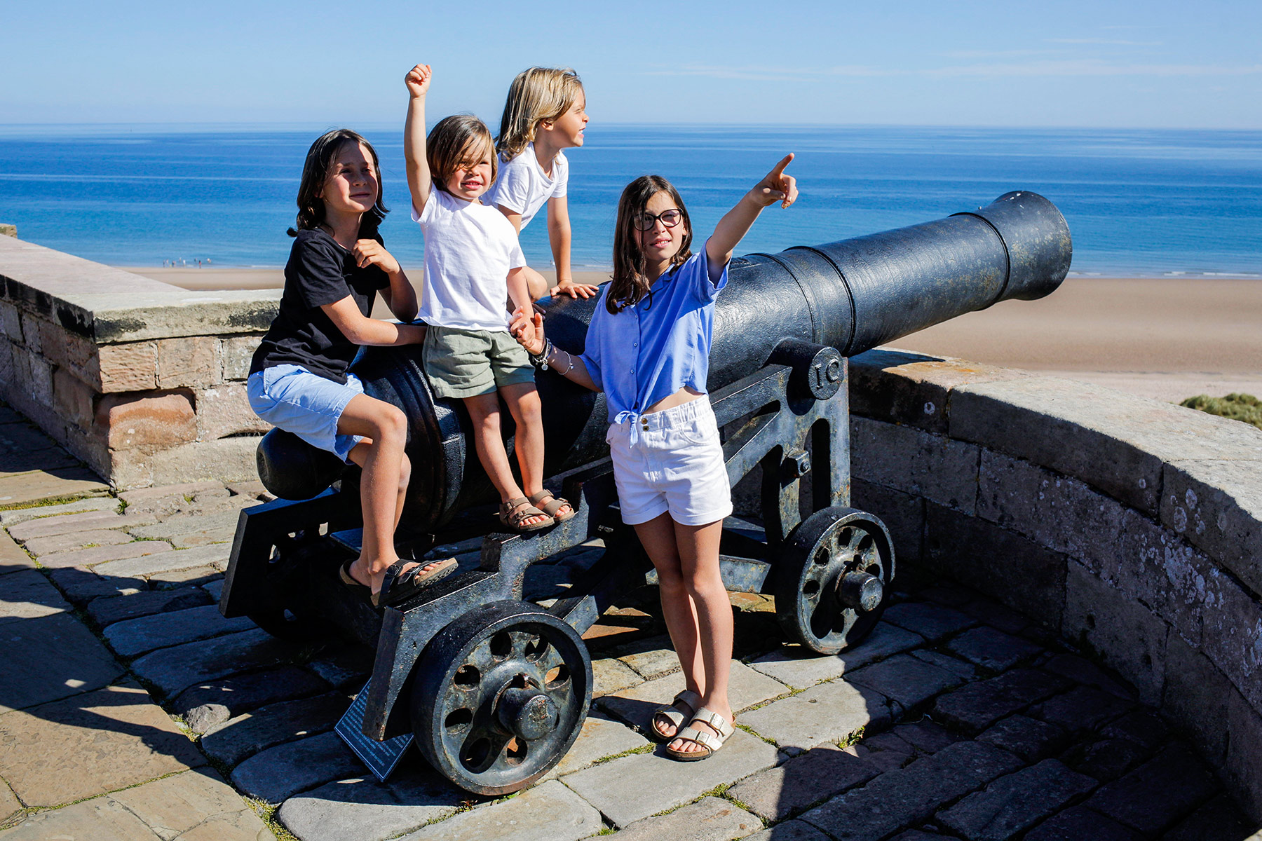 A group of children sit on a cannon on a castle wall with a blue sea and sand behind them.