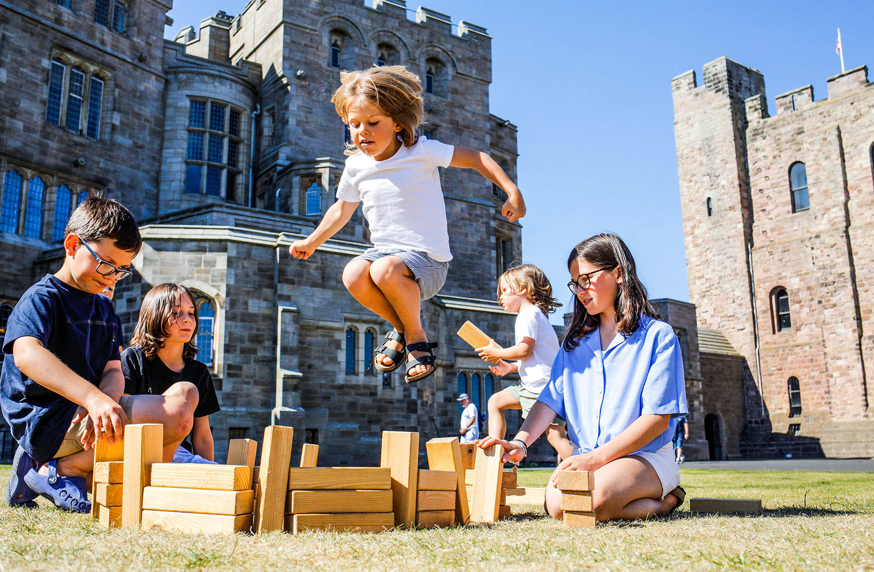 Children play with wooden blocks outdoors in front of a castle wall. One child is leaping over a small tower of the wooden blocks.