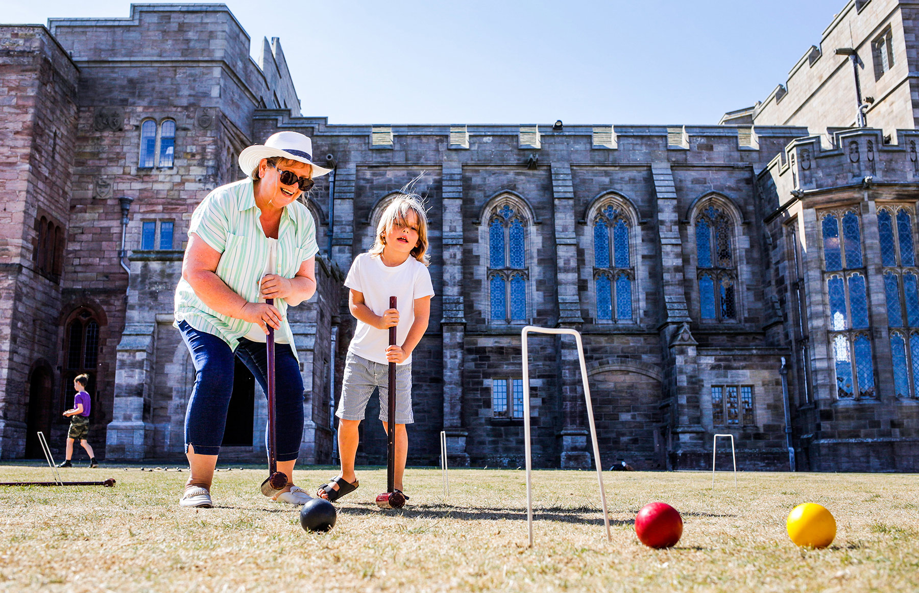 A child and grandparent play croquet on a front lawn in front of an old grand building.