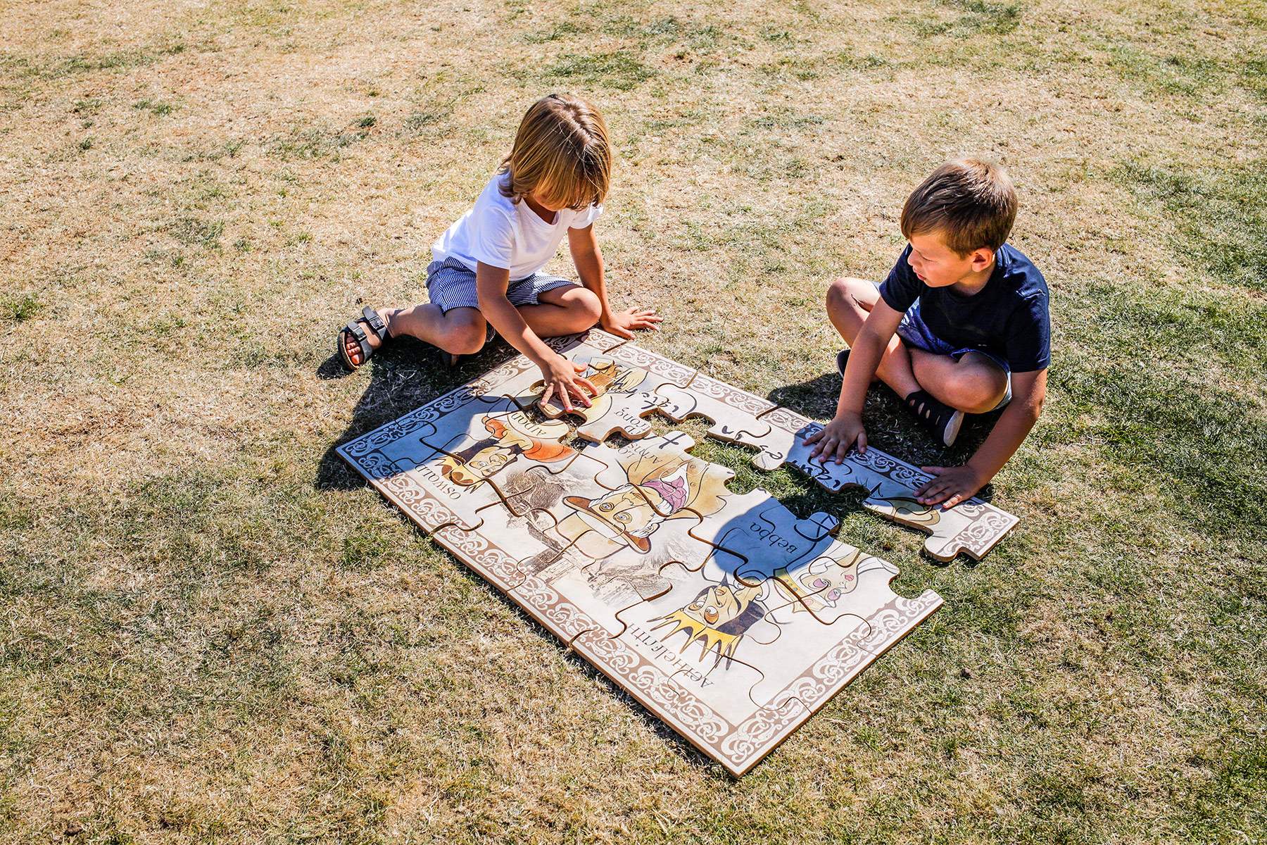 Two children sit on a grassy lawn putting together a large wooden jigsaw that shows kings and queens.