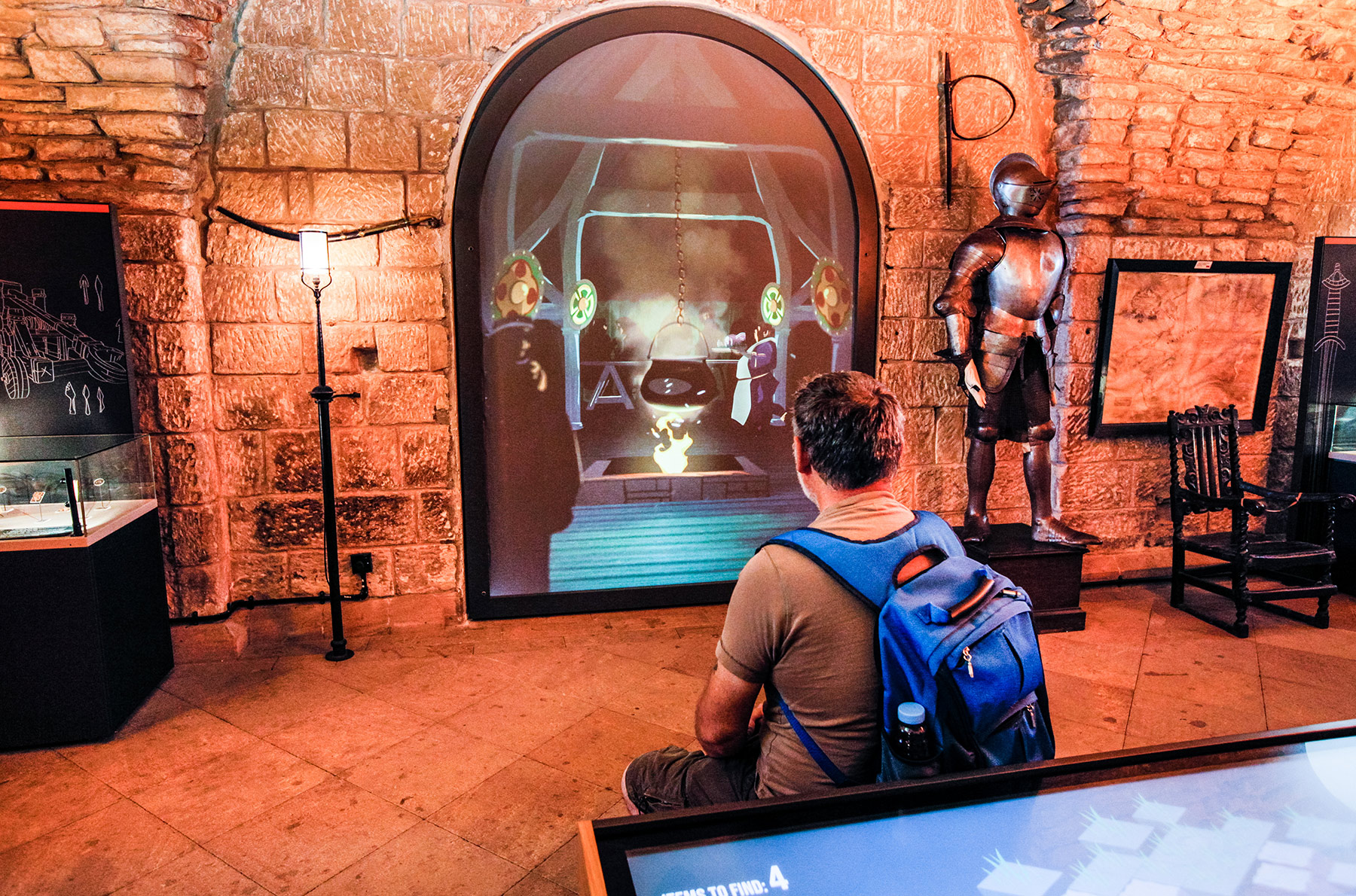 A man sits with a blue backpack on, in front of an interactive screen in an old part of a castle. There is a knight in armour to the right hand side of the screen.