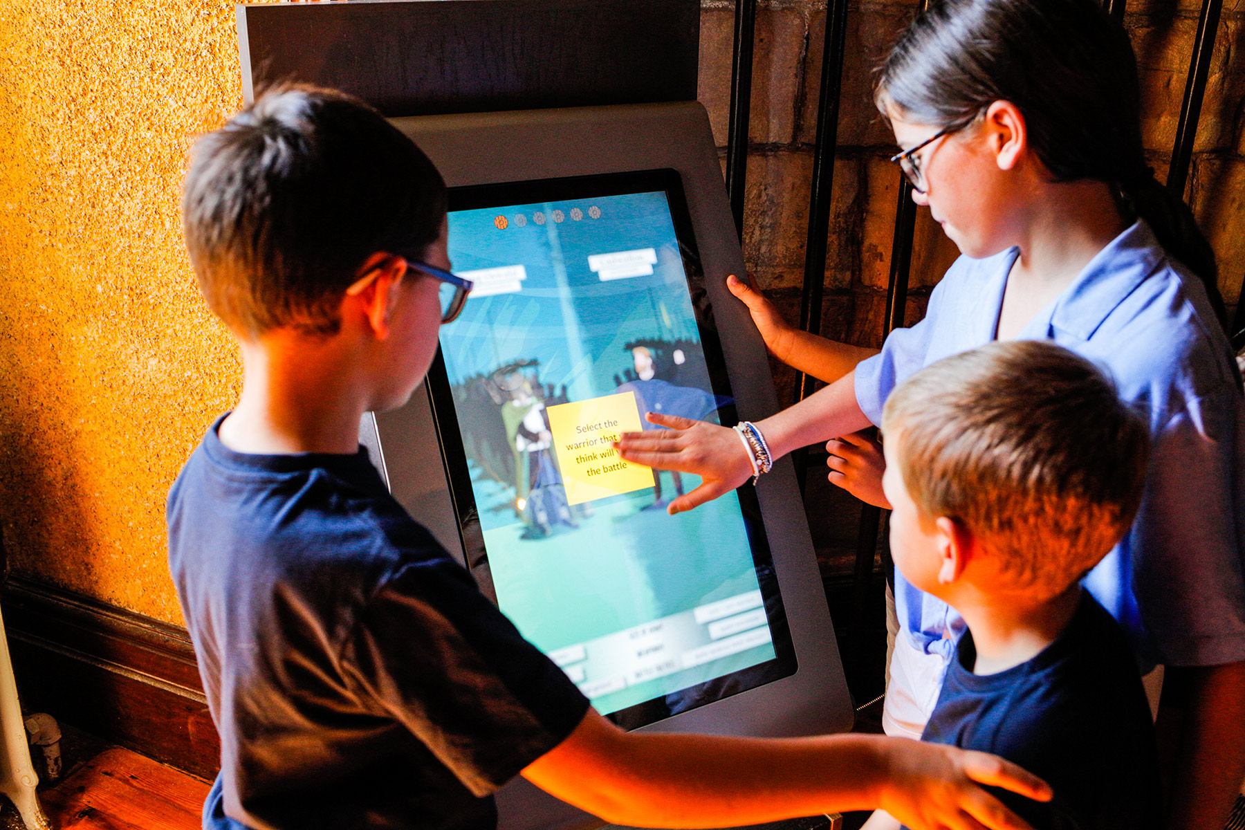 Three children look at an interactive screen and one of them is pushing a yellow button with writing on the screen.