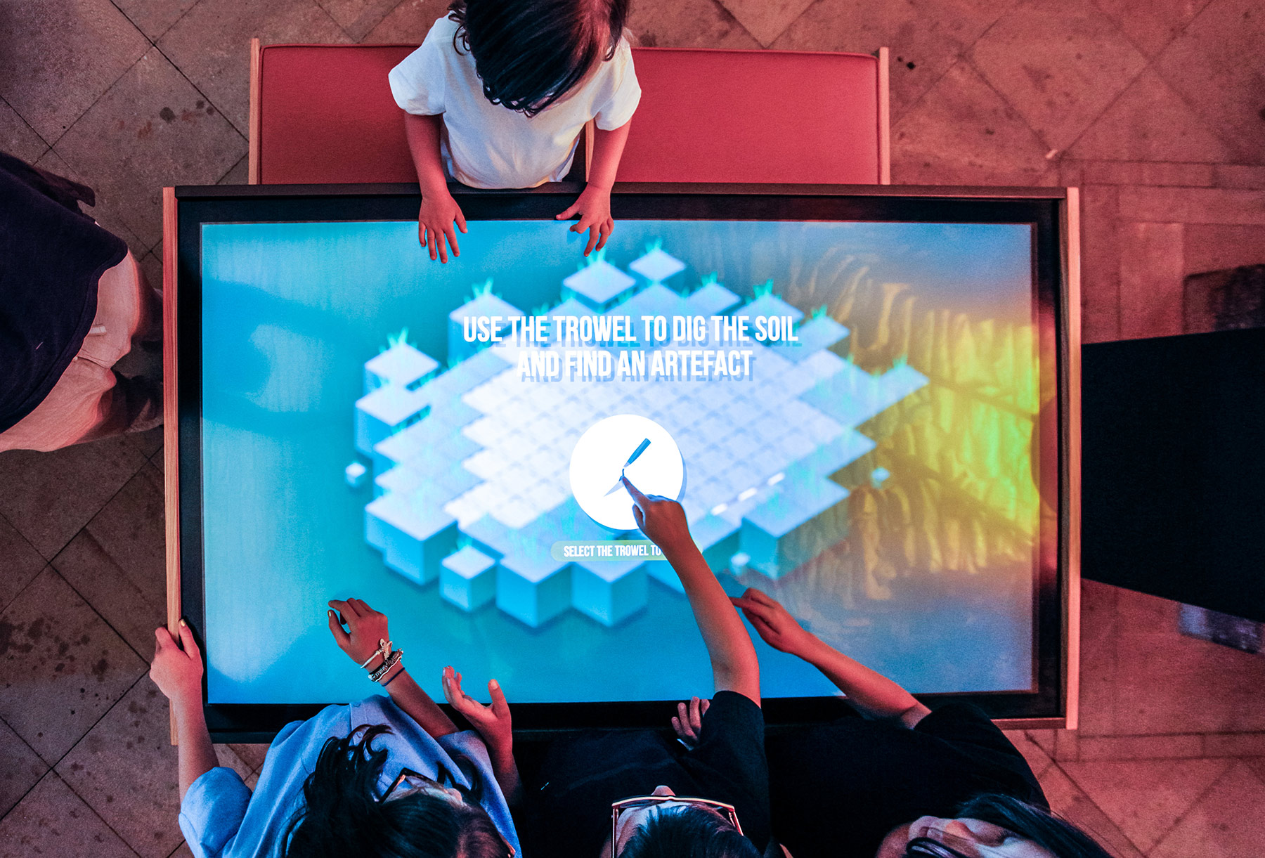Children stand around an interactive screen and touching buttons in the centre.