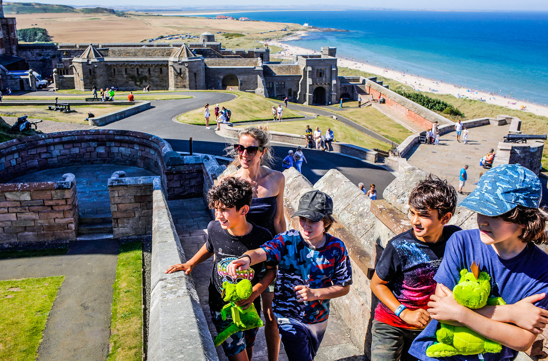 A woman with four boys climb castle steps. In the background the sea is vivid blue and people are dotted on the beach.