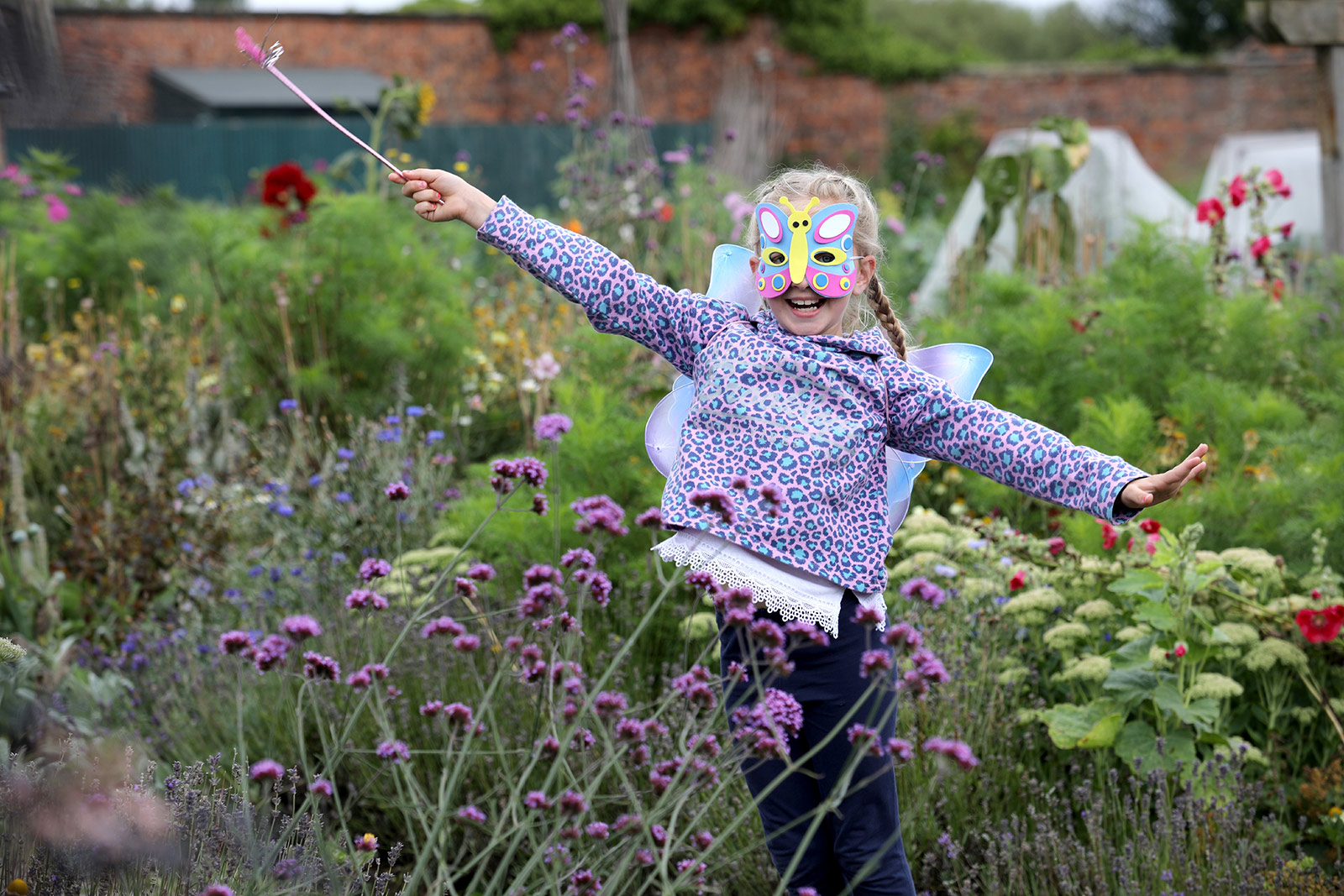 A young girl stands among flowers wearing a butterfly mask and holding a fairy wand.