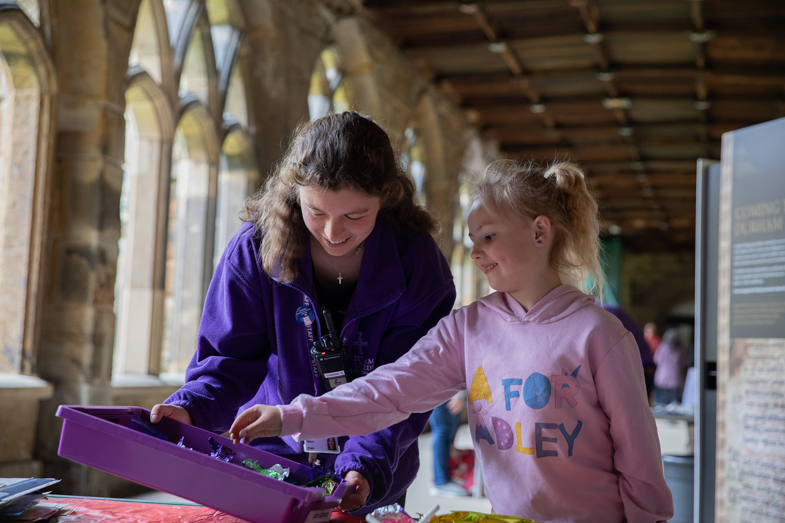 3Q8A9725_8inch A team member from Durham Cathedral is wearing a purple jumper and lanyard. She is holding up a purple tray from which a young girl take out an object.