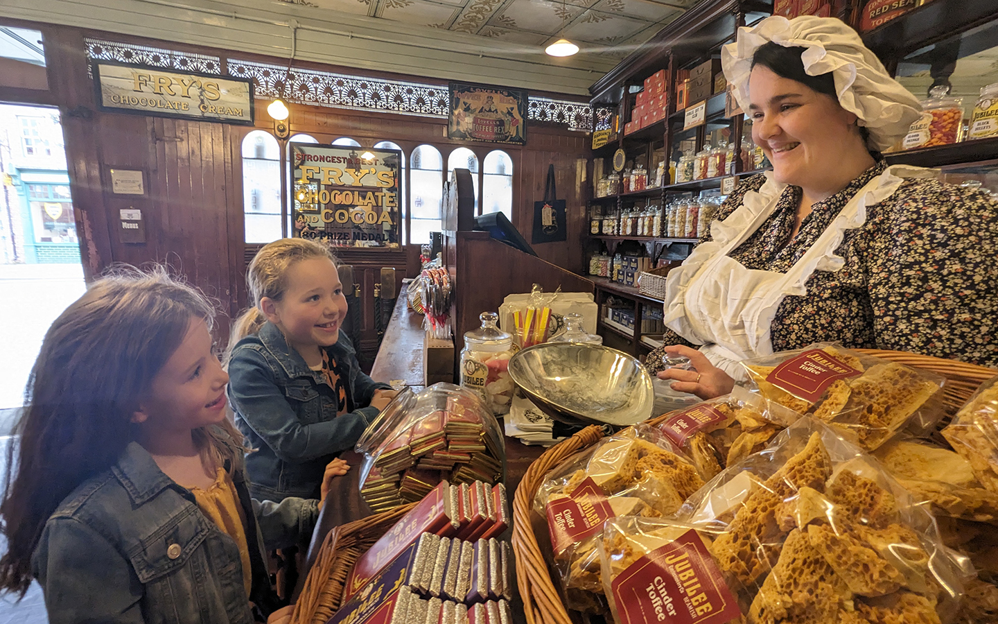 Beamish Museum, sweet shop Two children smile at a woman dressed in 1950s dress. They are in an old sweet shop with jars of traditional sweets on the shelves.