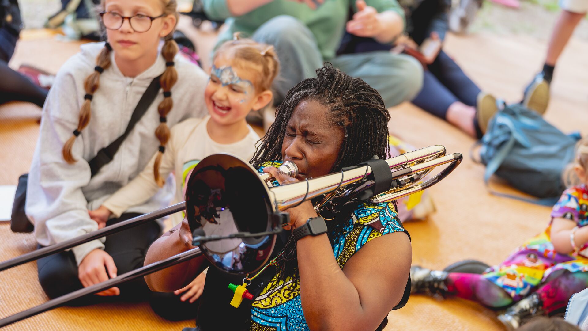 An adult blows into a trombone with young children laughing in the background