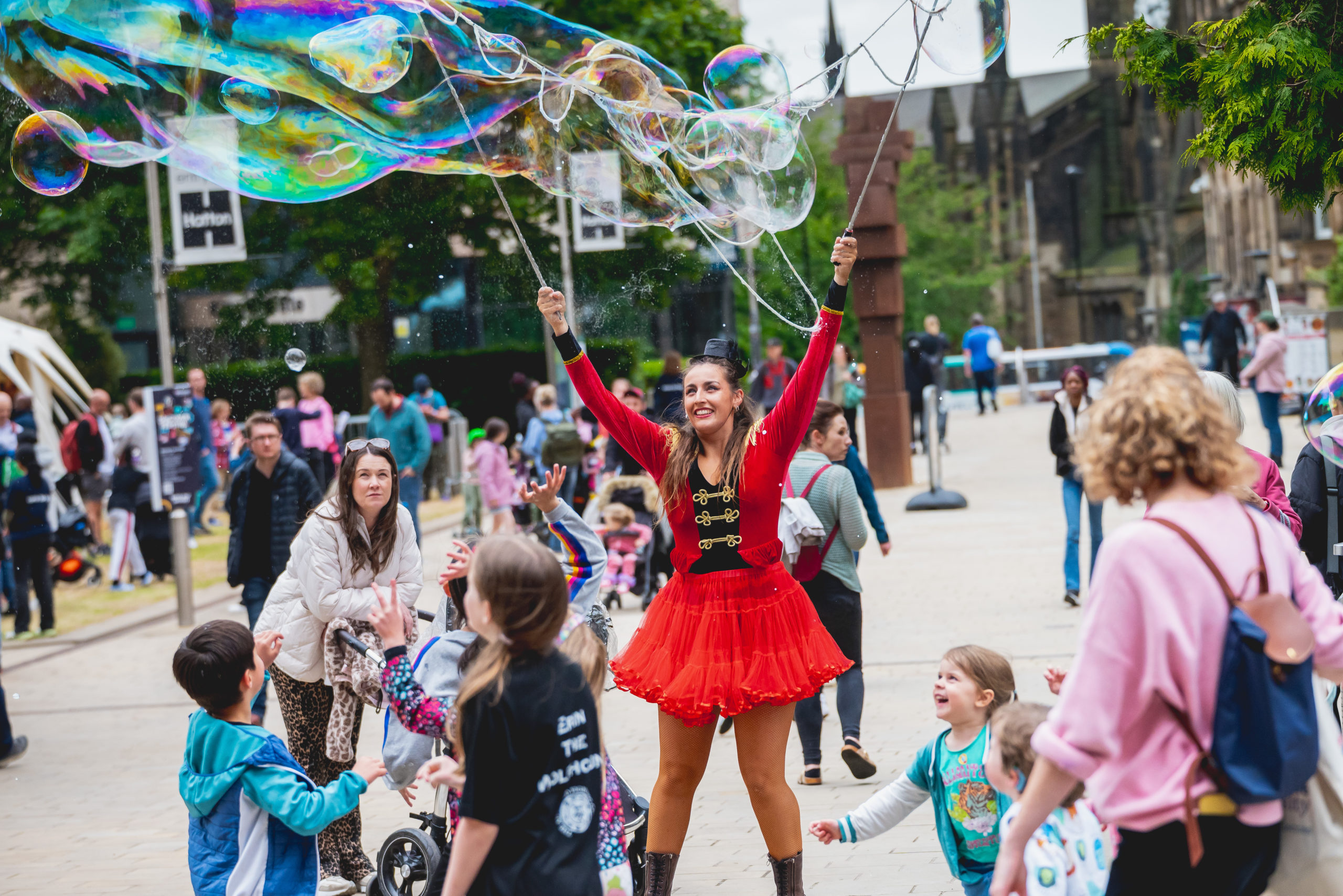 An adult in a bright red costume makes enormous bubbles as an excited crowd of families look on.