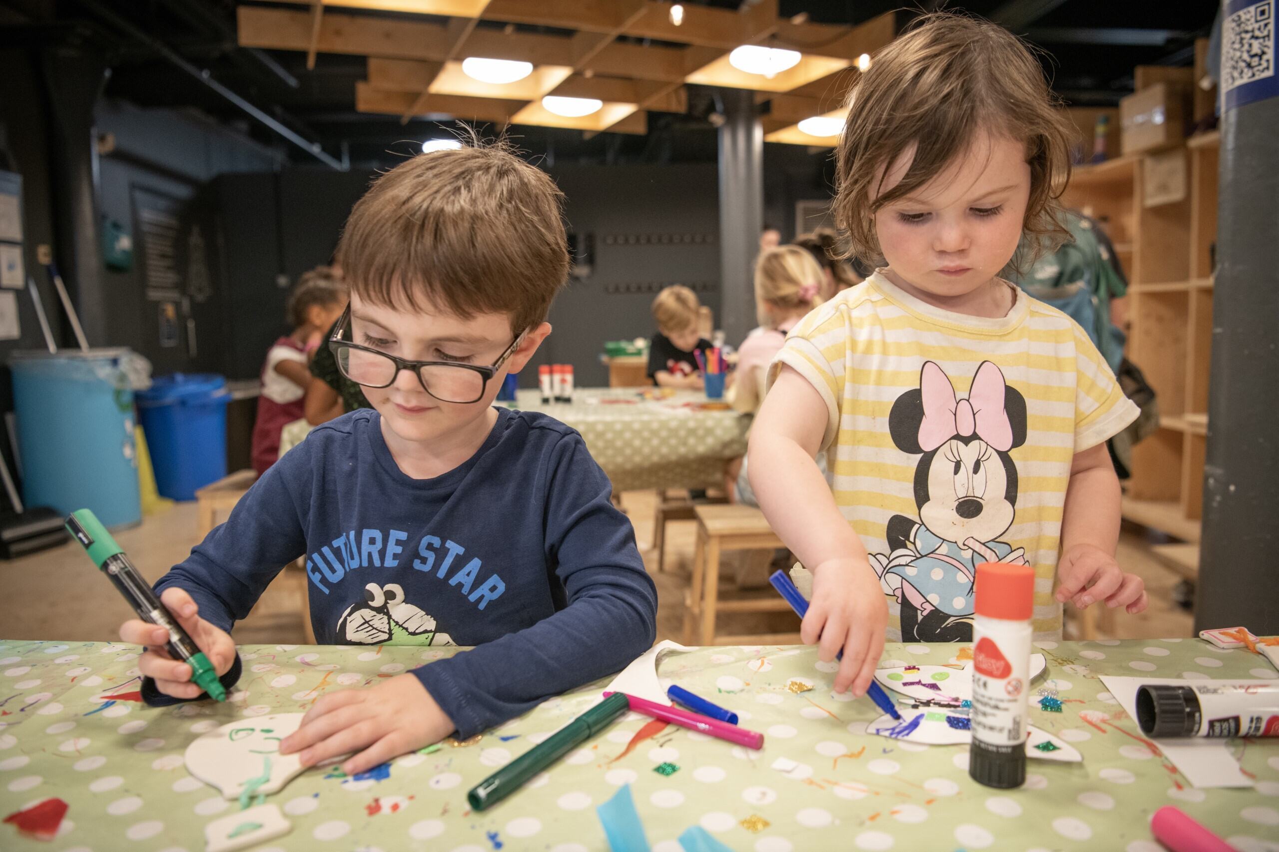 Discovery Museum 2 A young boy and girl are at a craft table holding coloured felt tips and drawing. Behind them are other children doing the same.