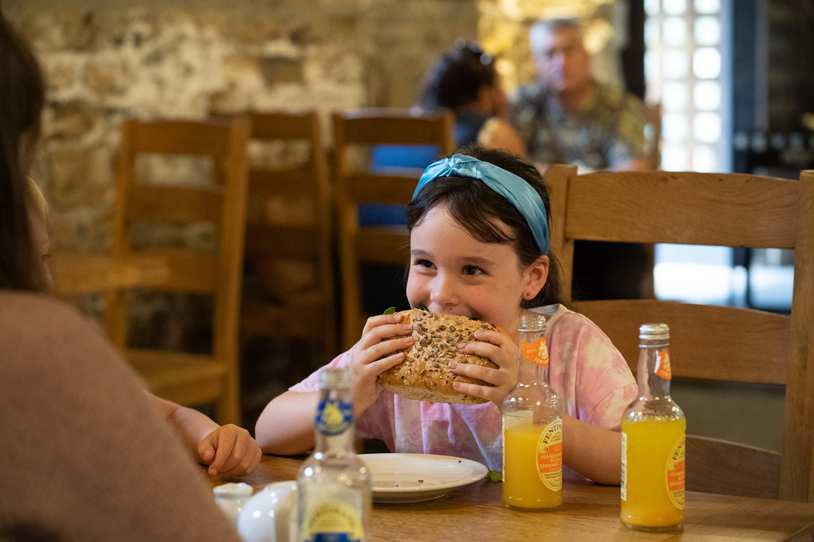 Durham-Cathedral-WEB-118 A young girl sits at the table in a cafe eating a large seeded bun. Drinks of orange juice are on the table.