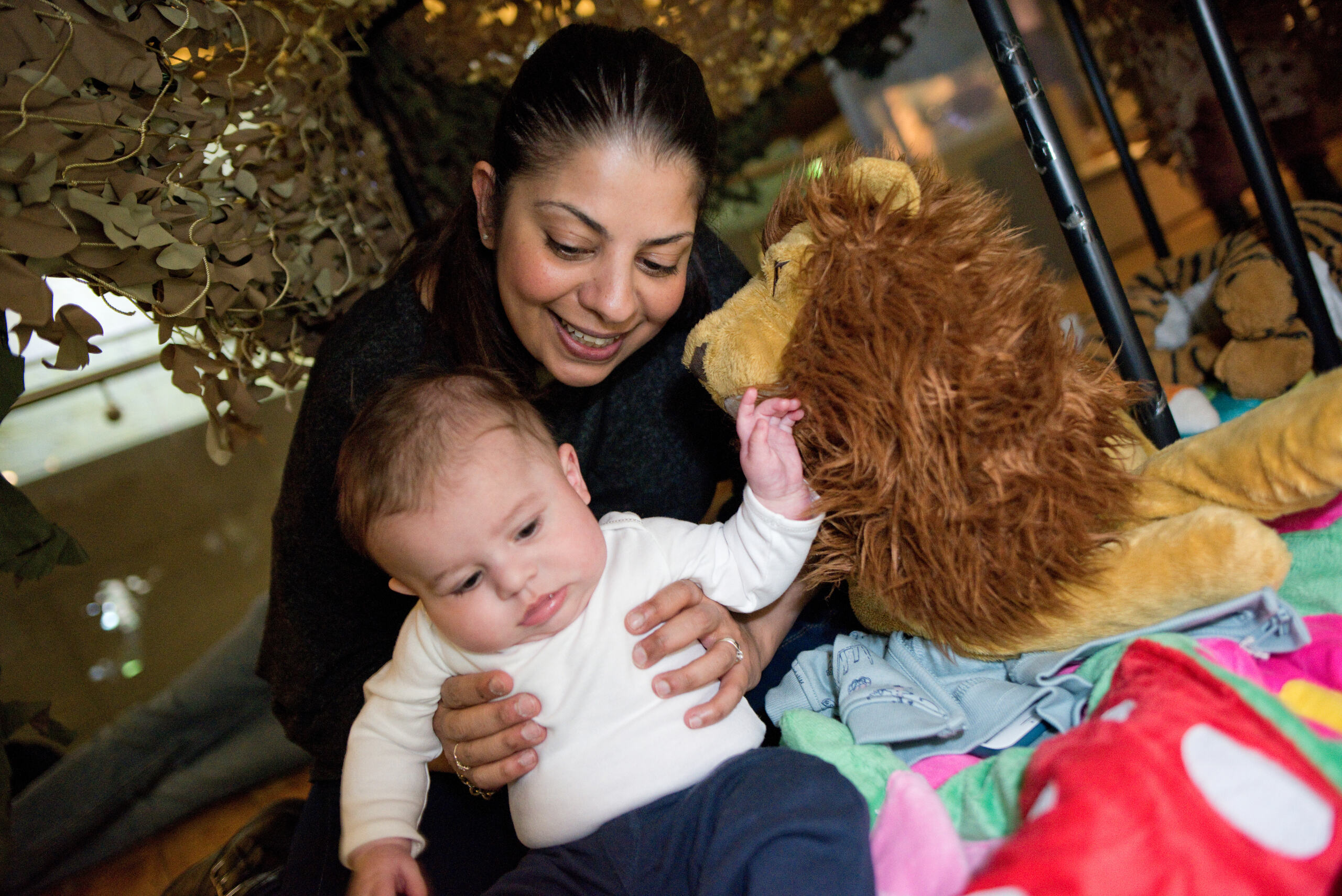 A mother holds her young baby as they sit surrounded by cuddly toys including a large lion.