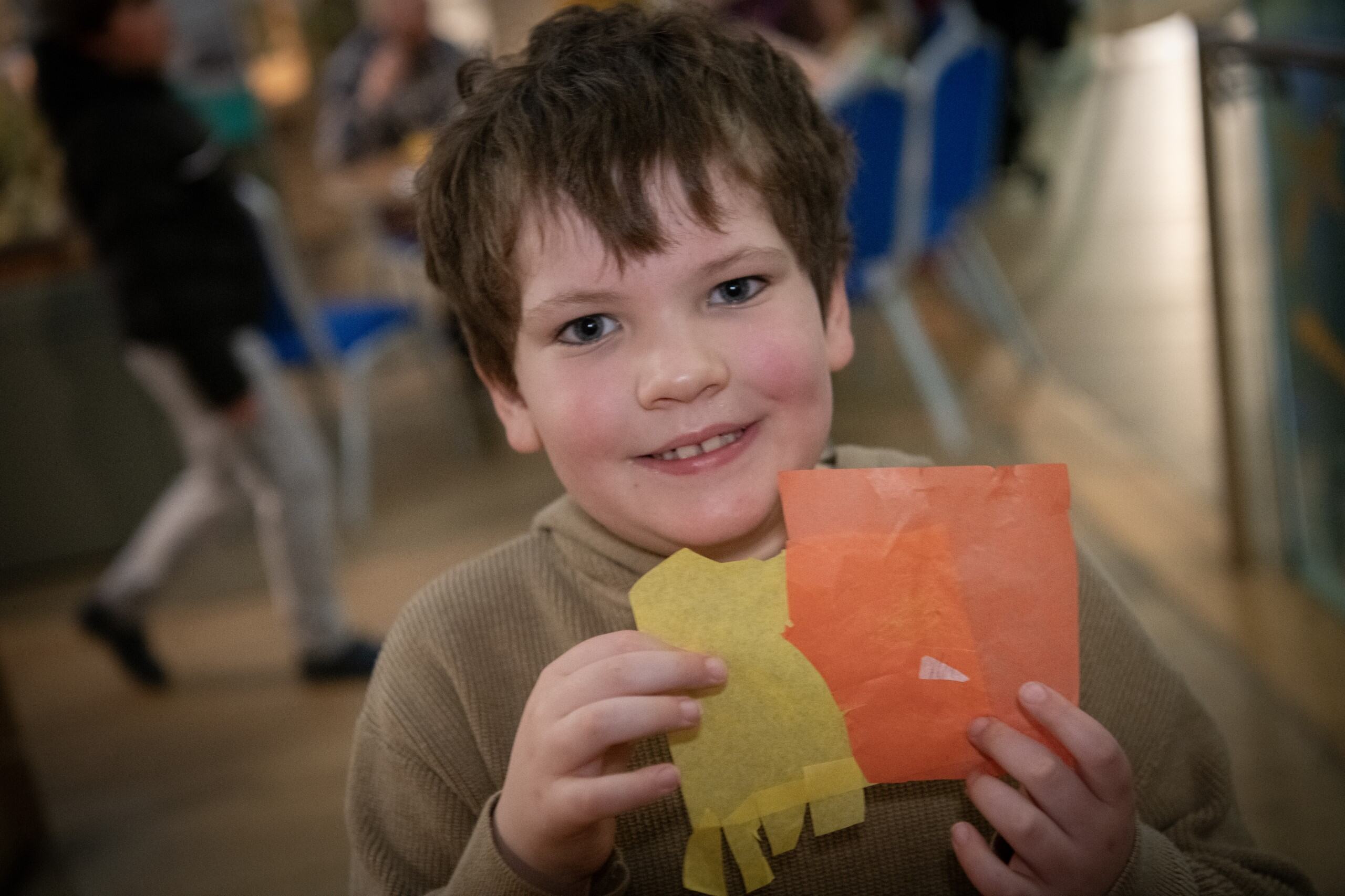 A young boy proudly holds in front of him a creation he has made from orange and yellow tissue paper.